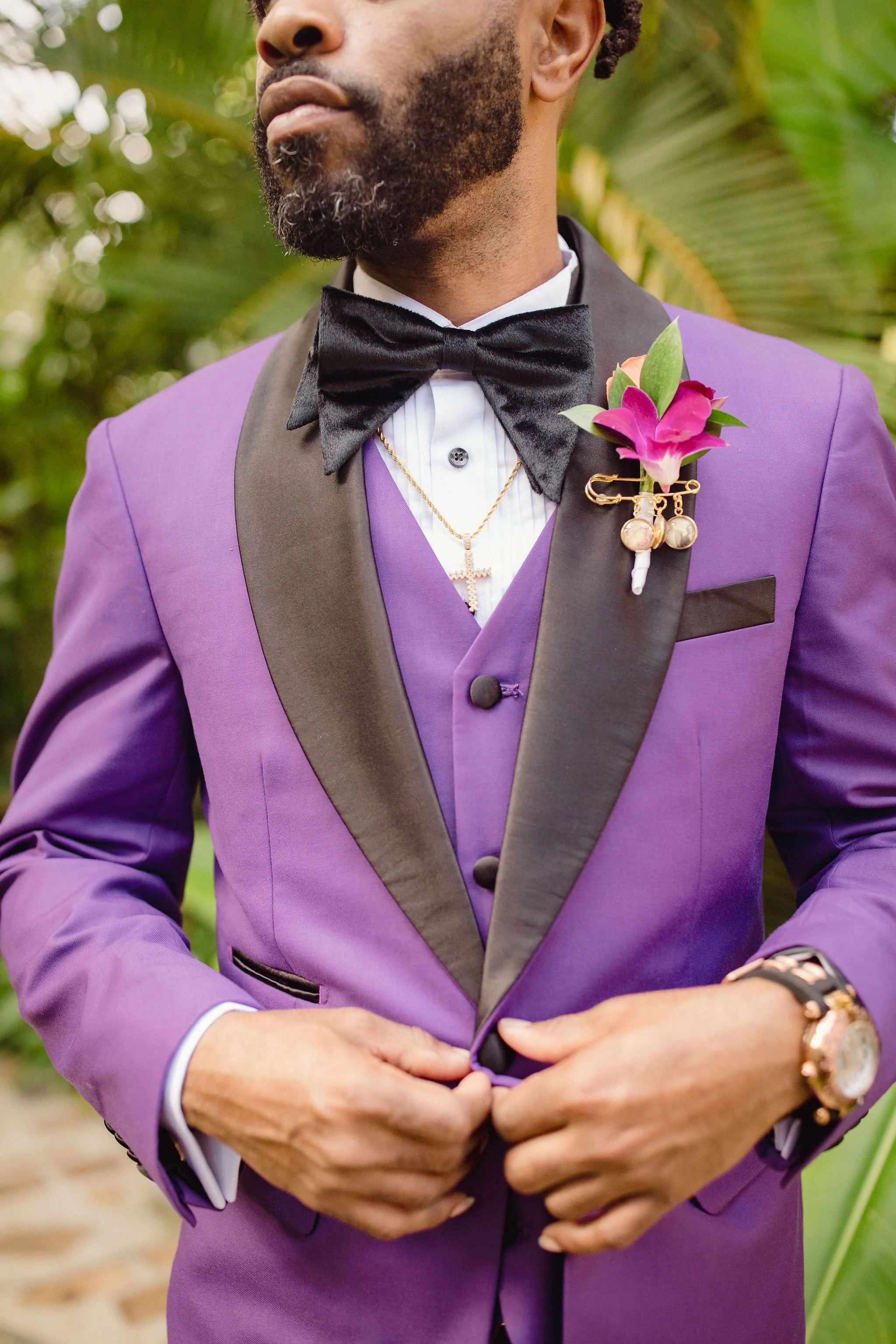 Close-up of a man in a purple tuxedo with black lapels, a black bow tie, and a boutonniere with a pink flower and decorative pins. He is adjusting his suit jacket.