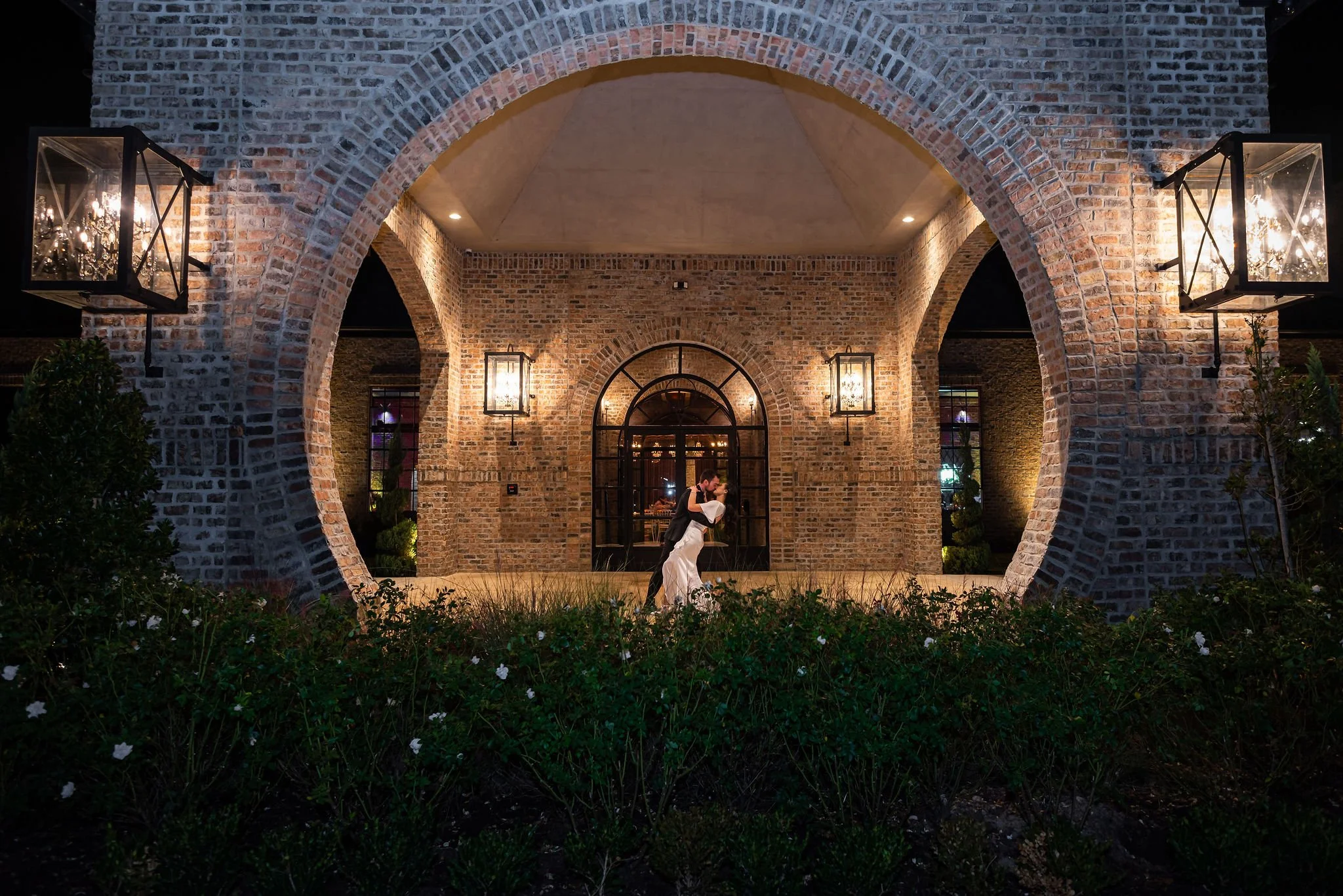 A bride and groom sharing a dance at night under a large brick archway with lanterns.