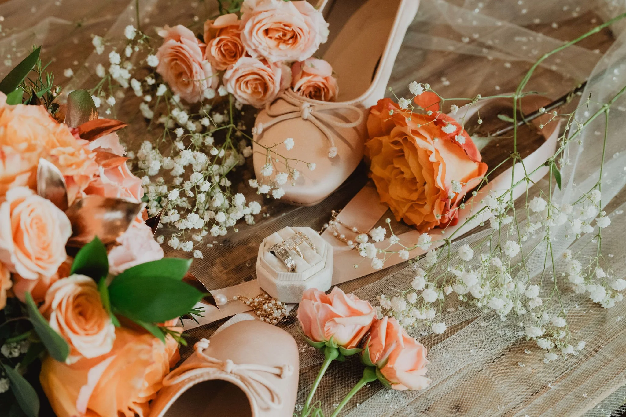 A wedding setup with peach and pink roses, baby's breath, wedding rings, and a ballet slipper on a rustic wooden table.