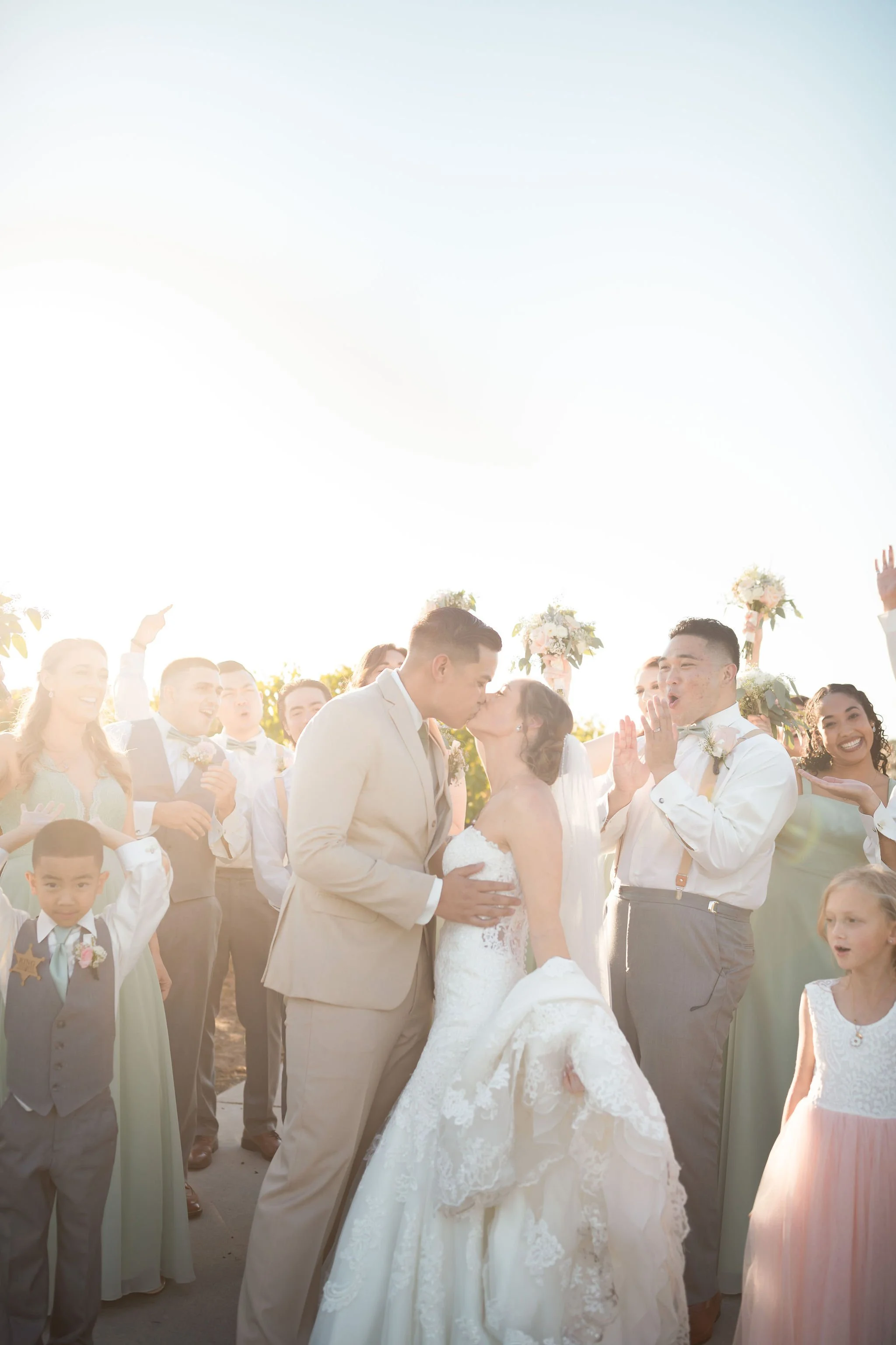 A wedding couple shares a kiss while surrounded by wedding guests, with the sunlight creating a bright, warm atmosphere.