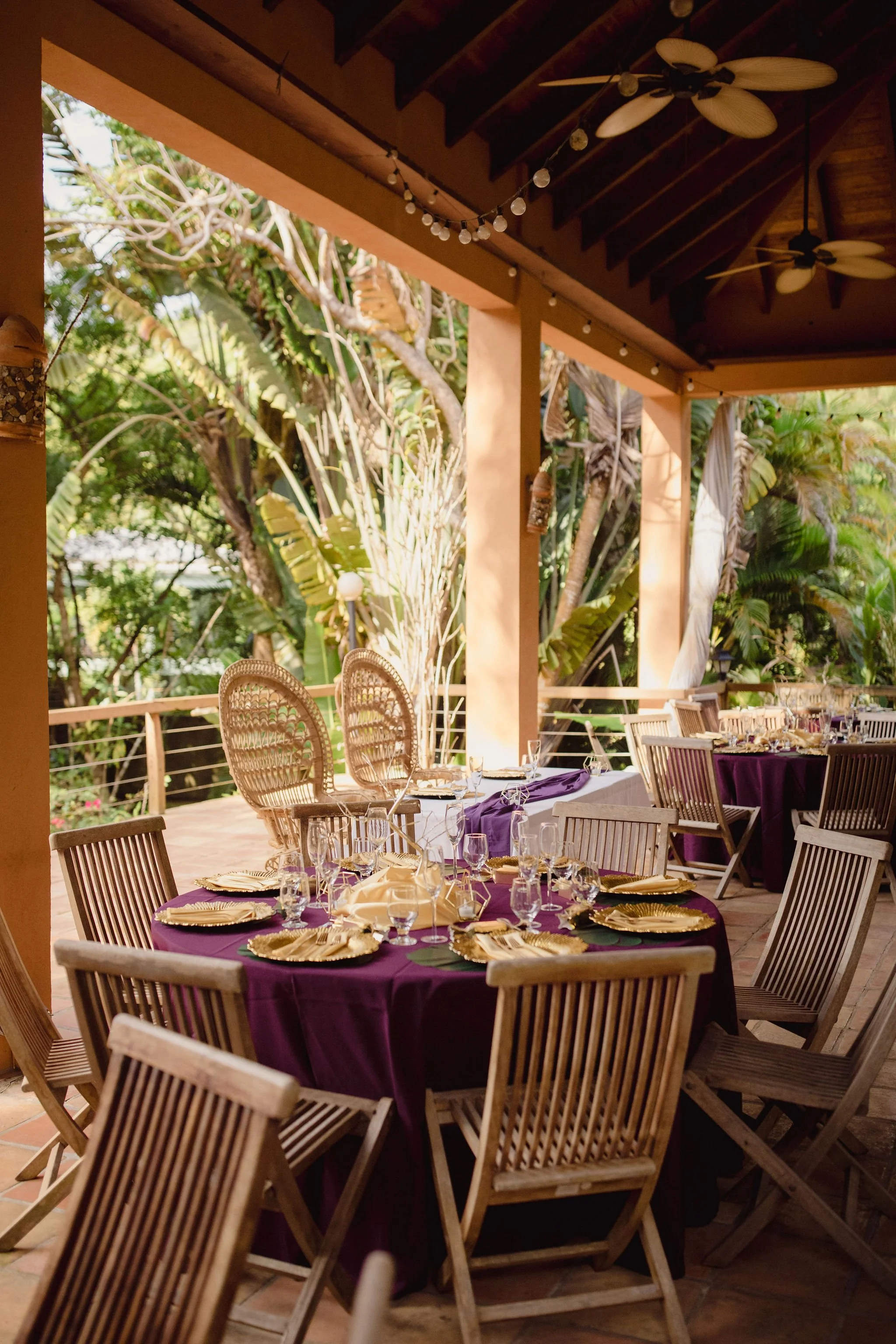 Outdoor dining area with round tables covered in purple tablecloths, set with gold plates, glassware, and utensils, surrounded by wooden chairs, with lush greenery in the background and string lights hanging from the ceiling.