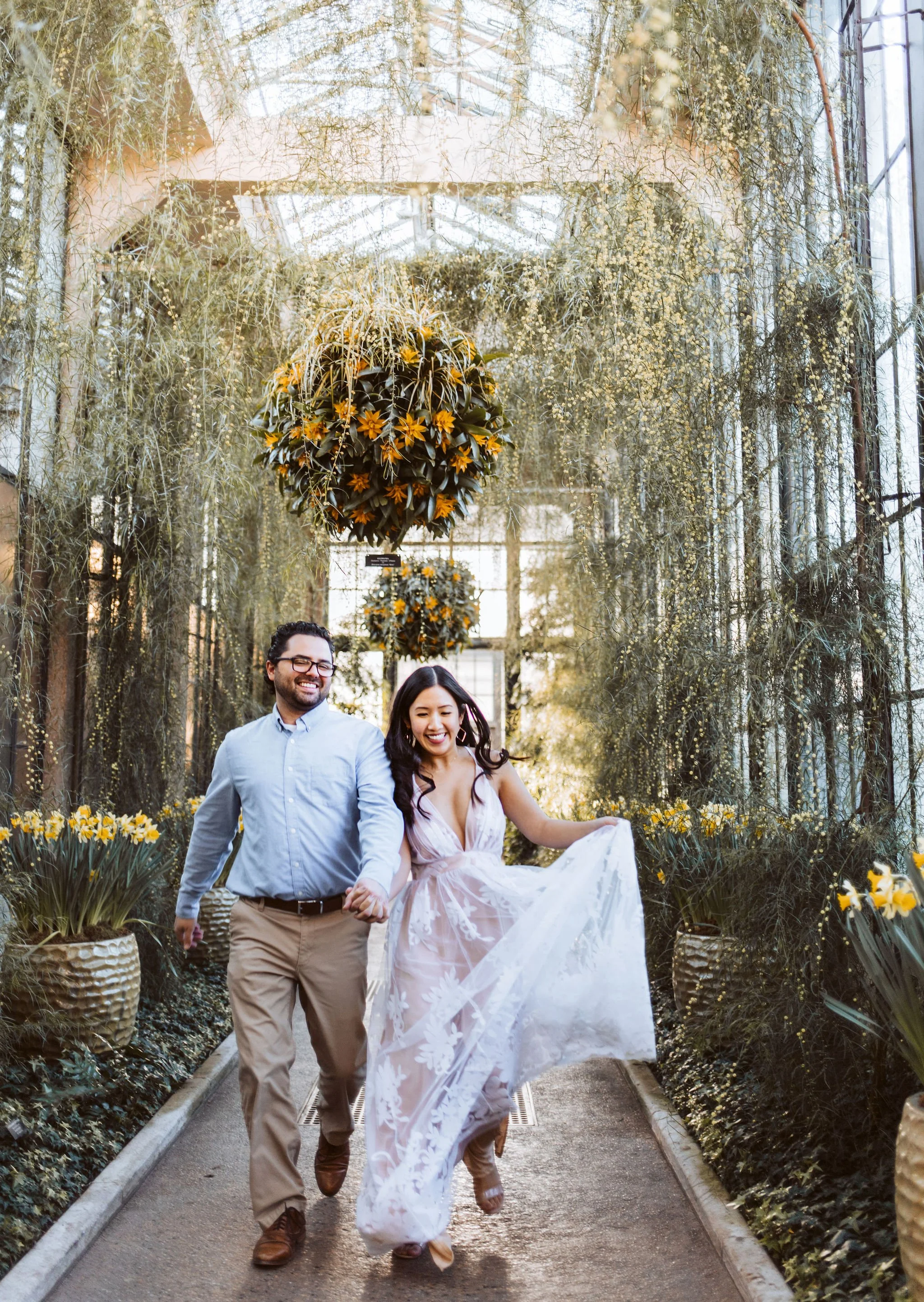A happy couple walking hand in hand through a lush, floral greenhouse filled with yellow flowers, natural greenery, and hanging plants on a sunny day.