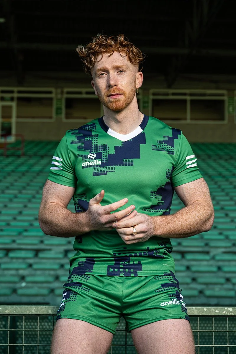 A young man with curly red hair and freckles, wearing a green and blue sports uniform with the logo 'O'neills,' standing in an empty stadium with green seating.