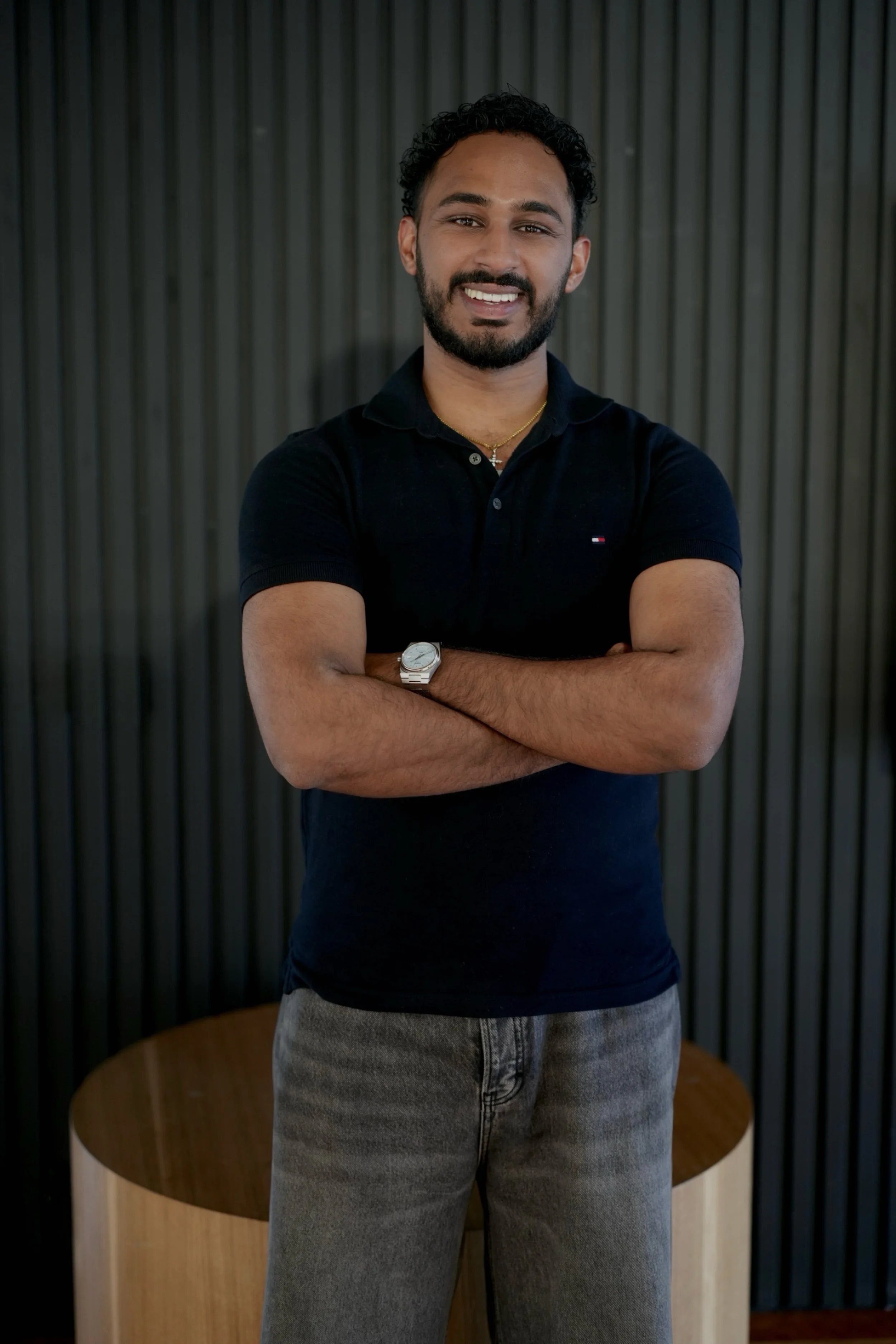 A man with dark curly hair, a beard, wearing a black polo shirt with a small logo, a gold necklace, a watch, and gray pants, standing with arms crossed in front of a dark background with vertical lines.
