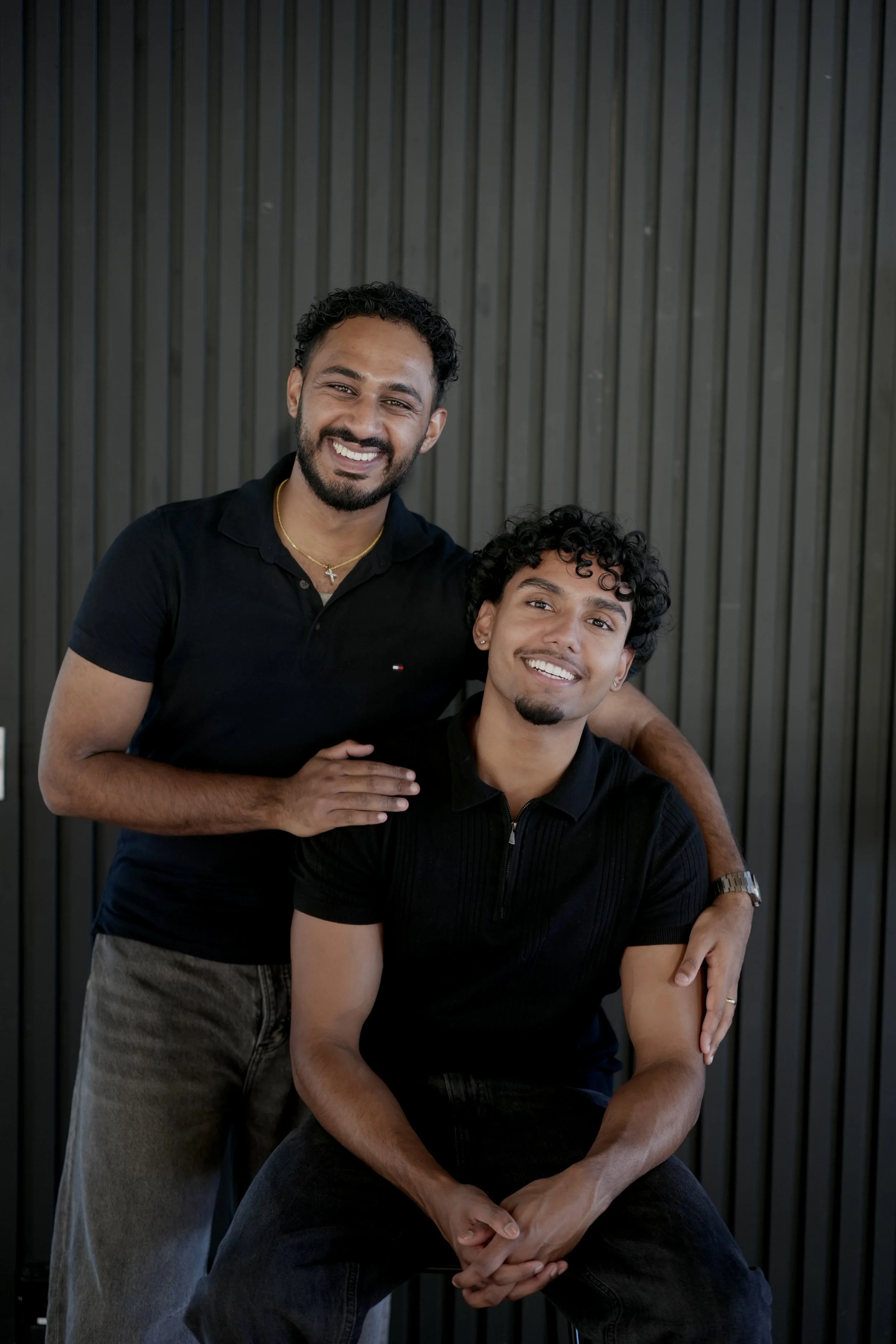 Two young men smiling and posing against a black, vertical wooden background. One is standing behind the other, with his arm around the seated man's shoulder.