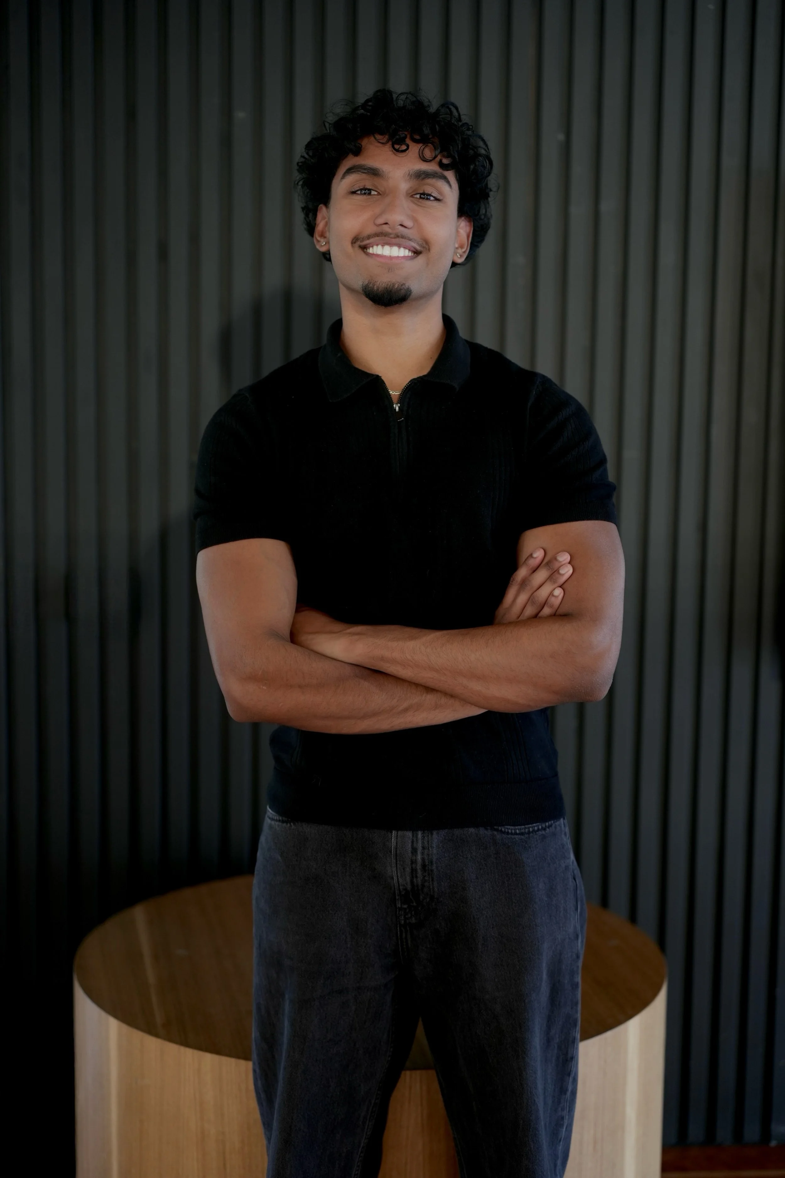 A young man with curly black hair, a goatee, and earrings smiling with arms crossed, standing in front of a dark, vertical-panel wall.