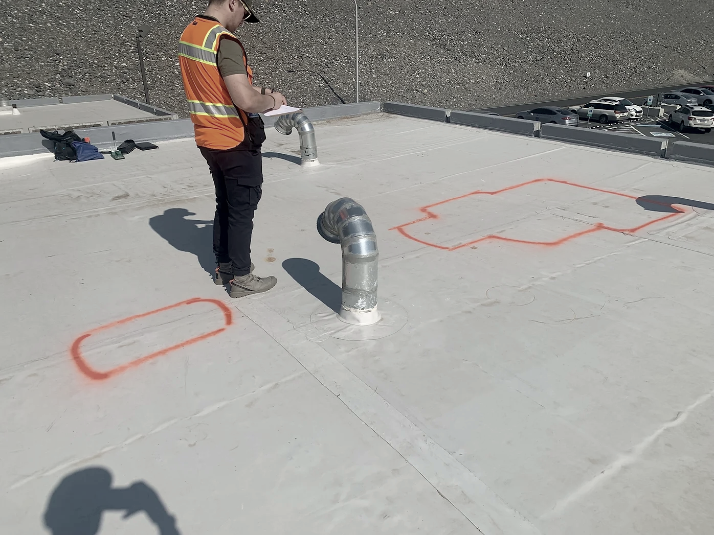 A person wearing a safety vest and helmet inspecting a flat rooftop with metal pipes and spray-painted markings in orange.