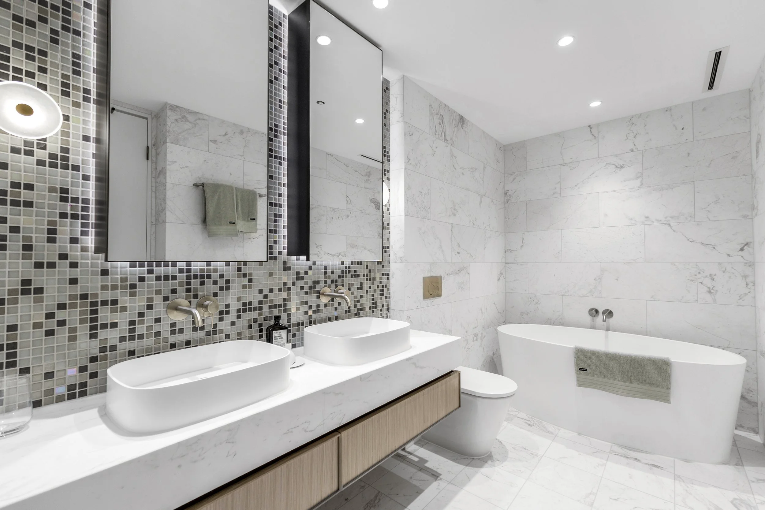 Modern bathroom with white marble walls and floor, a double sink vanity with vessel sinks, a large mirror, a bathtub with a towel, and modern lighting.