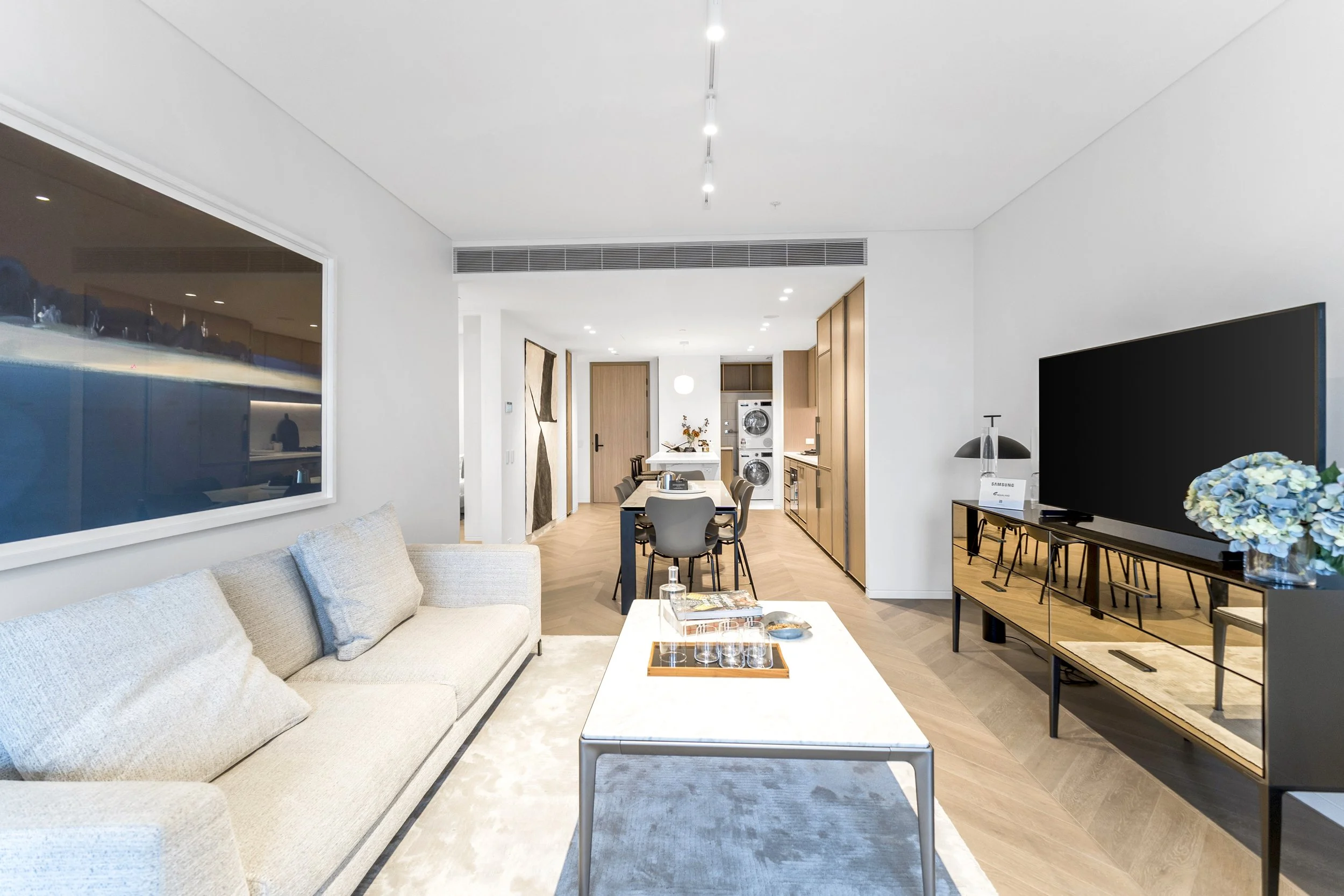 Modern living room with beige sofa, white coffee table, flat-screen TV, and dining area in the background