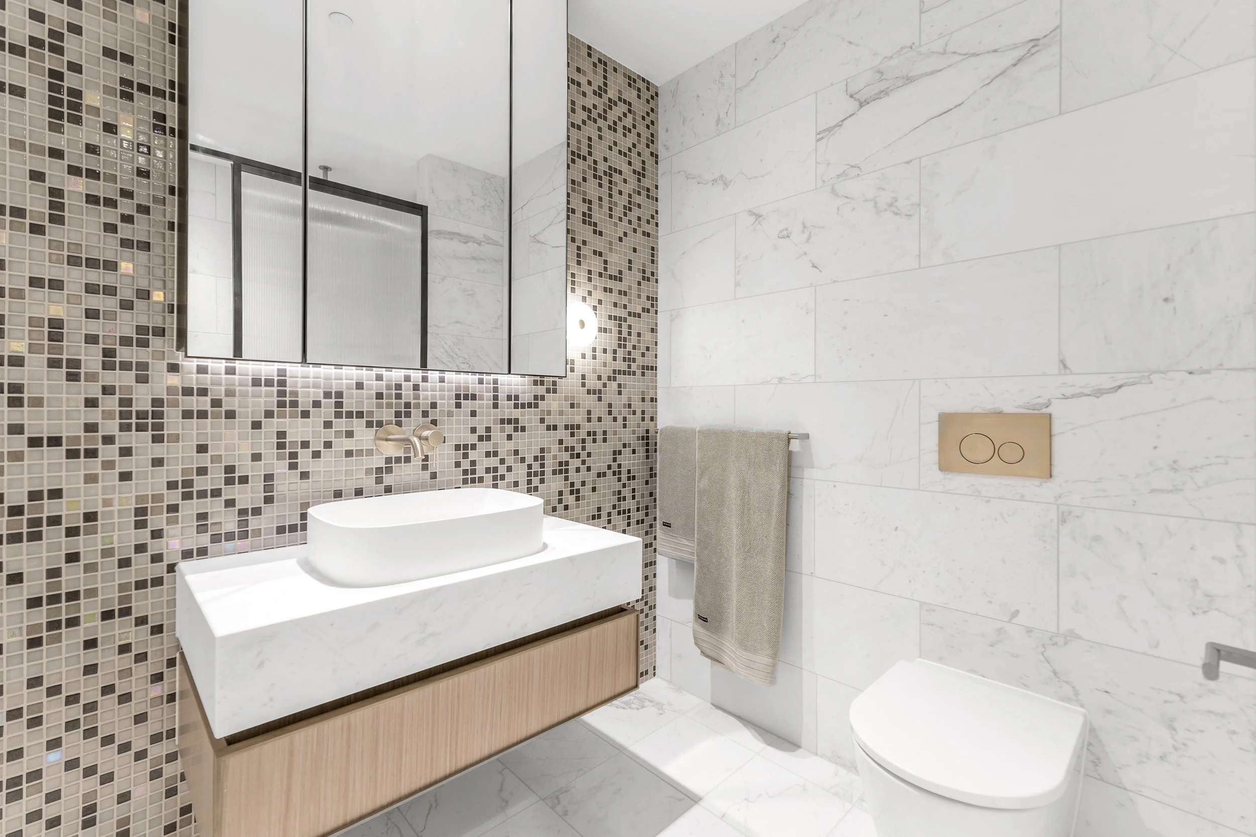 Modern bathroom with white marble walls and floor, mosaic accent wall, large mirror, vessel sink, towel rack with beige towel, and concealed toilet flush panel.