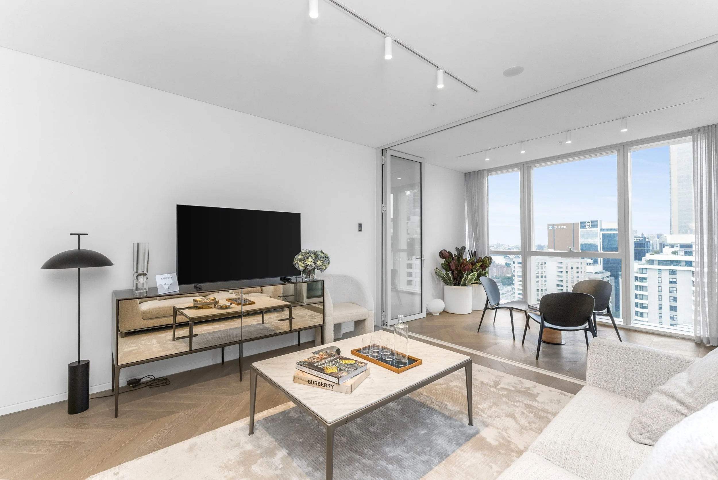 Modern living room with large windows, beige sofa, marble coffee table with books and glassware, black chairs near window, TV on wooden sideboard, and potted plants.