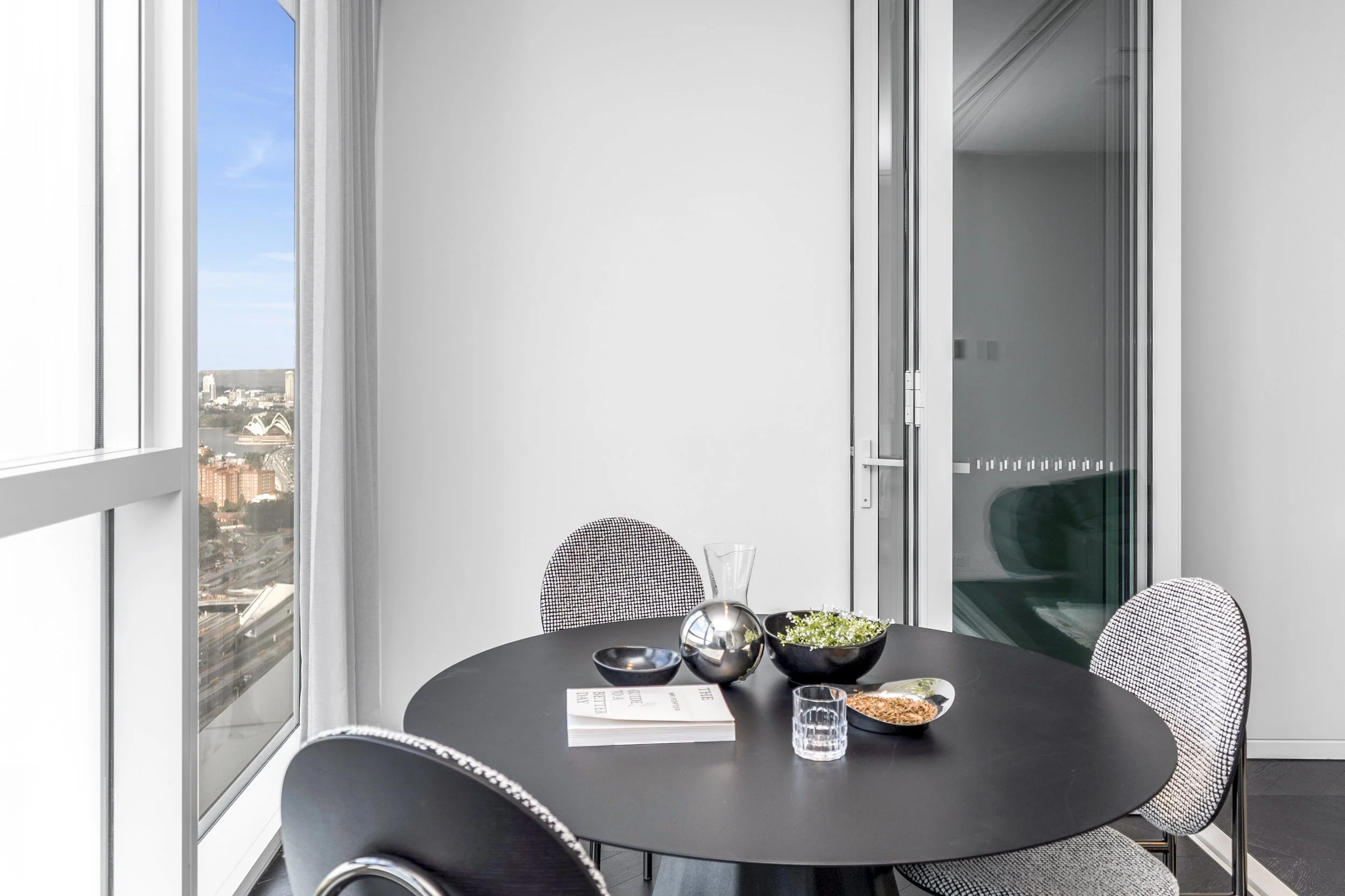 Modern dining area with a black round table, patterned chairs, and decorative items, with large windows showing a cityscape including a recognizable Sydney Opera House.