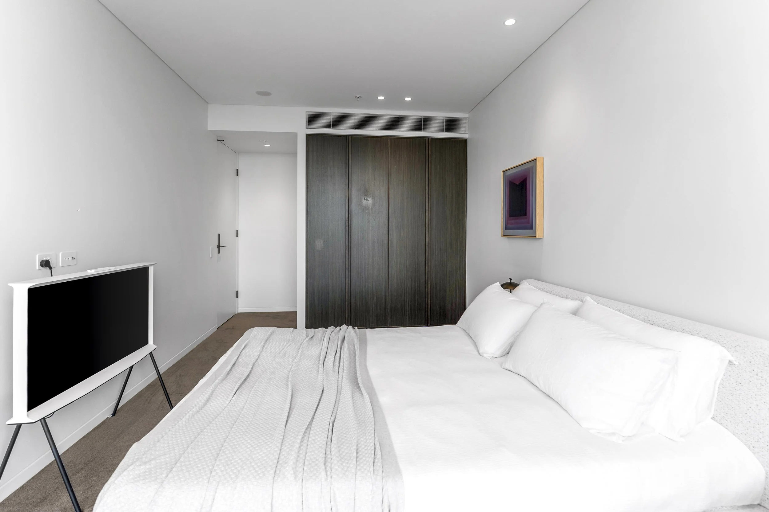 A modern hotel room with a white bed, a flat-screen TV on a stand, a framed artwork on the wall, and dark closet doors.