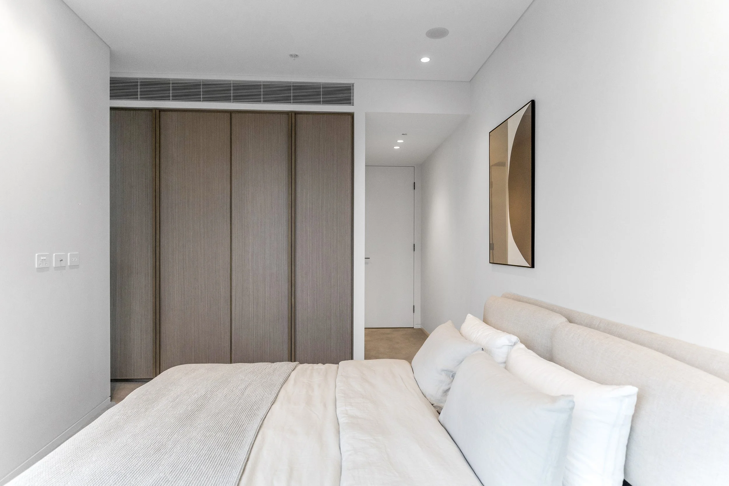 A bedroom with a beige bed, white pillows, a white wall, a wooden closet, and a modern abstract art piece on the wall.