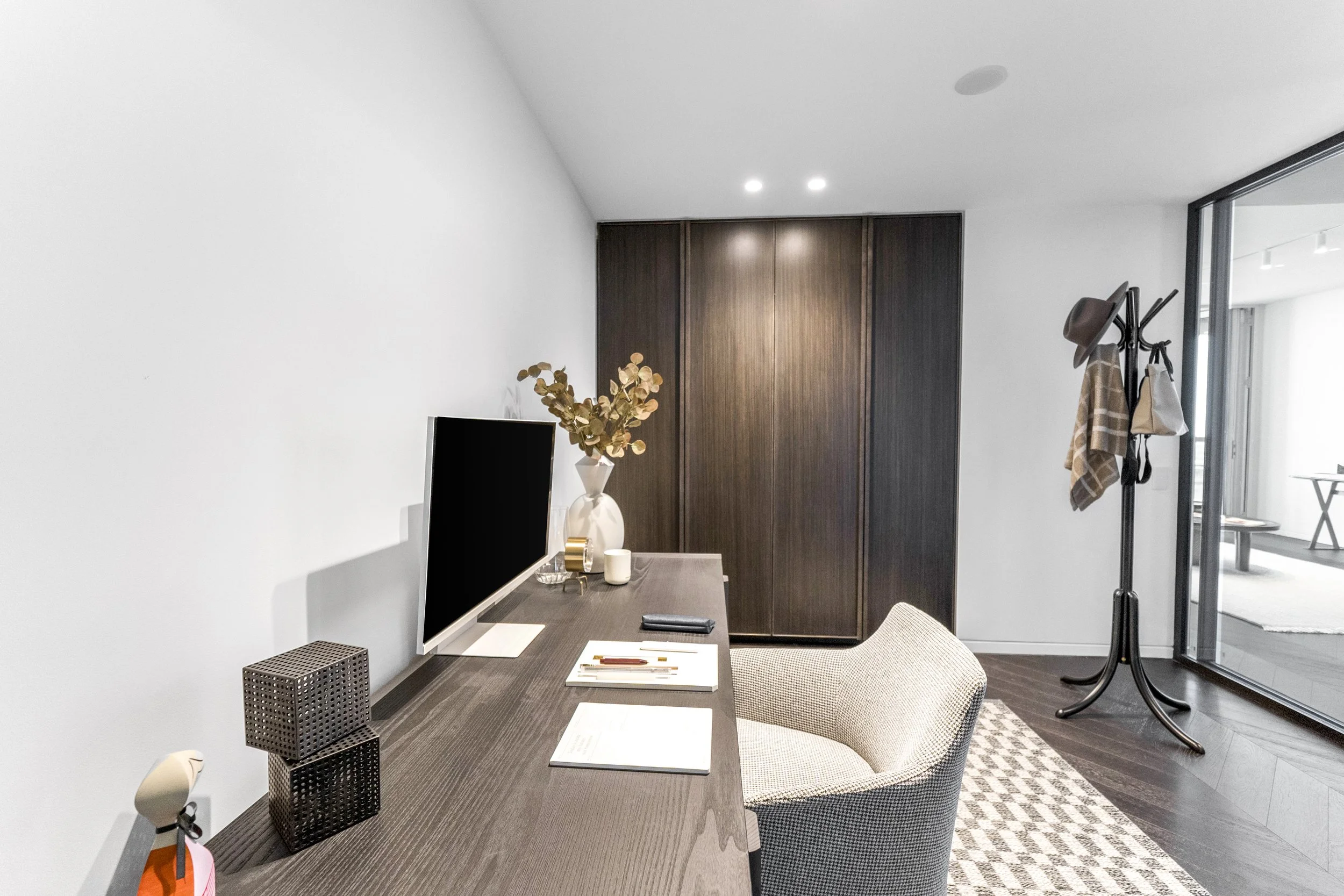 Modern home office with wooden desk, desktop monitor, vase with dry flowers, lamp, notebooks, and a chair, adjacent to a coat rack with hats, scarves, and bags near a sliding glass door leading outside.