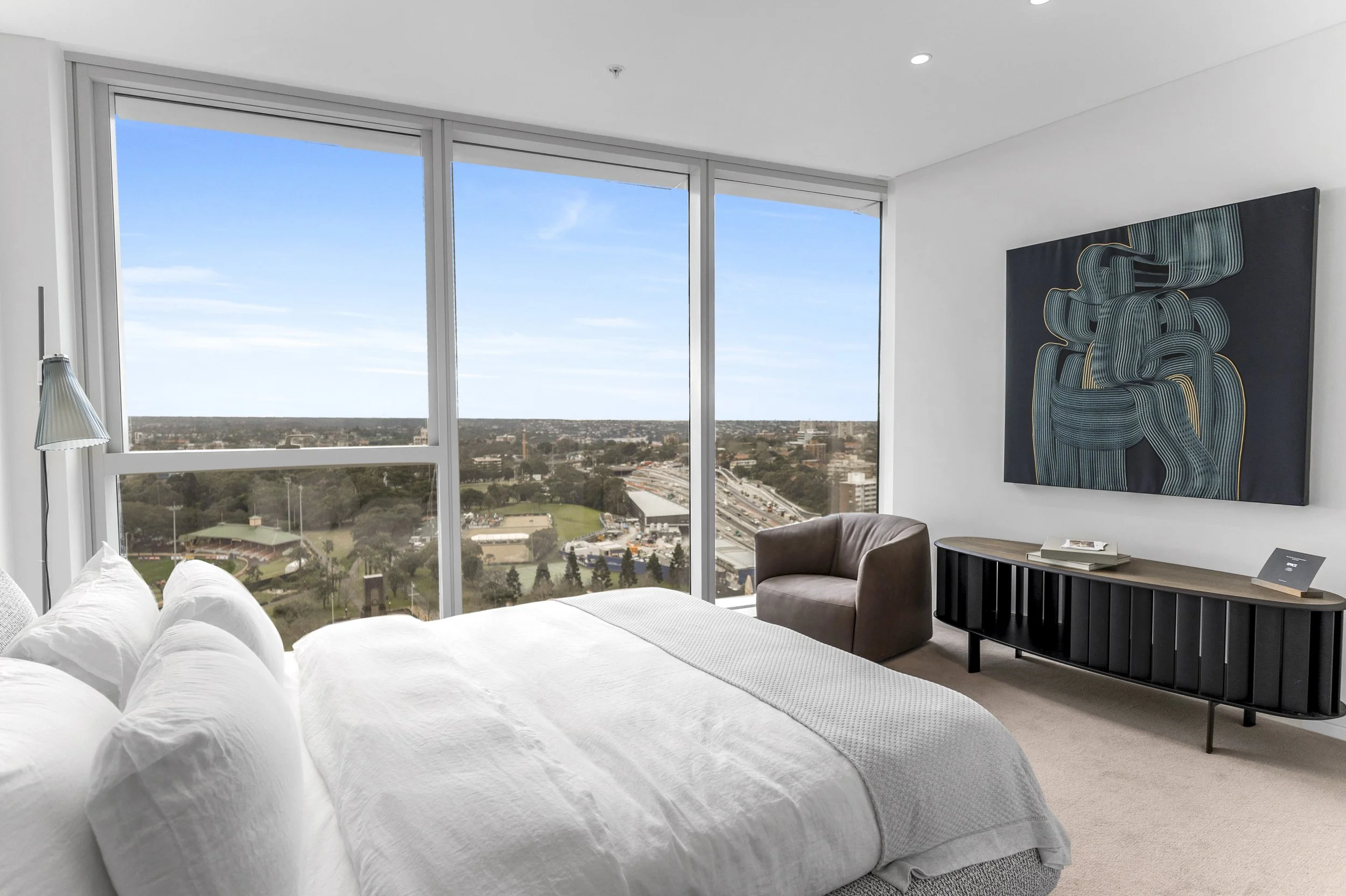 Modern bedroom with large windows showing city view, white bed, brown armchair, black console table, abstract artwork on the wall.