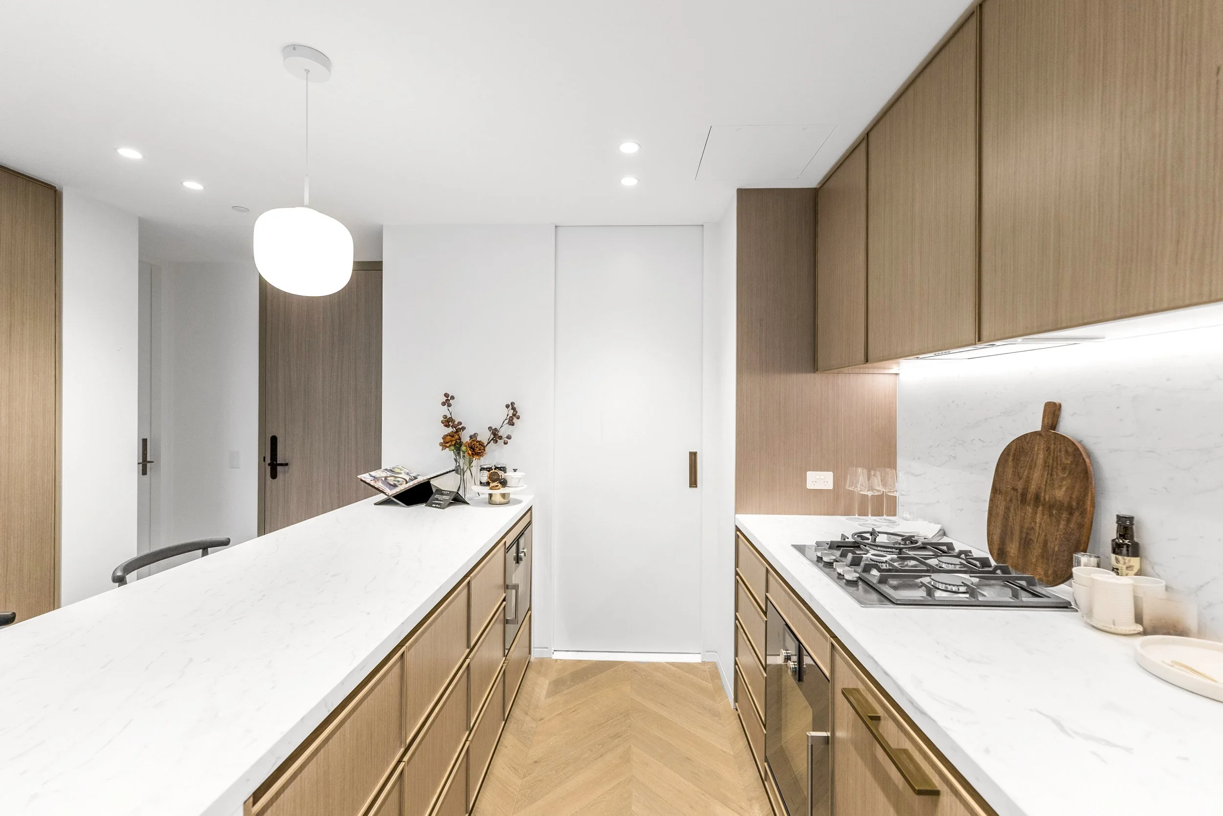 Modern kitchen with white marble countertops, wooden cabinets, gas stove, cutting board, wine glasses, and decorative items.