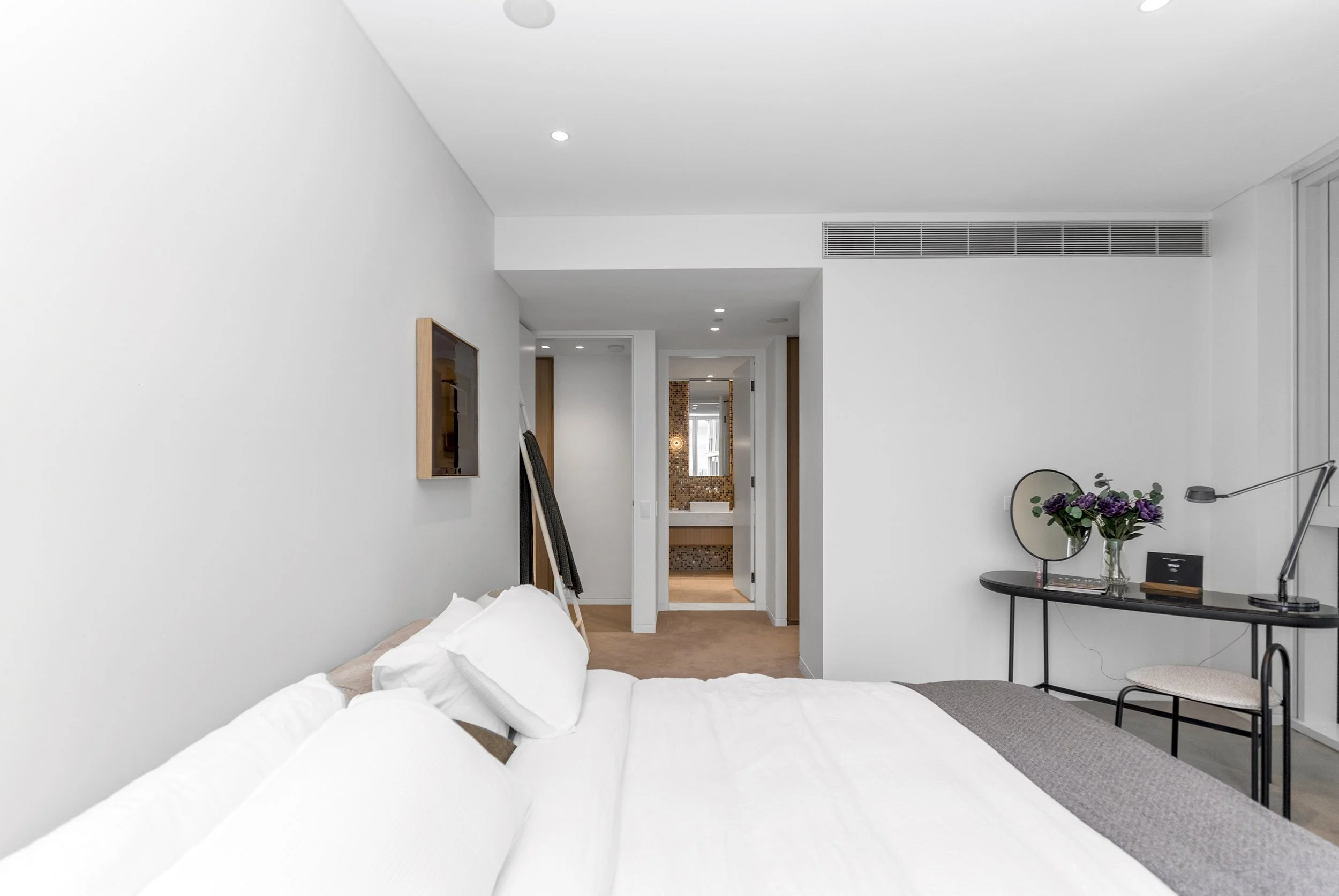Hotel room with a bed, a small desk with a mirror, a vase of flowers, and an open bathroom door in the background.
