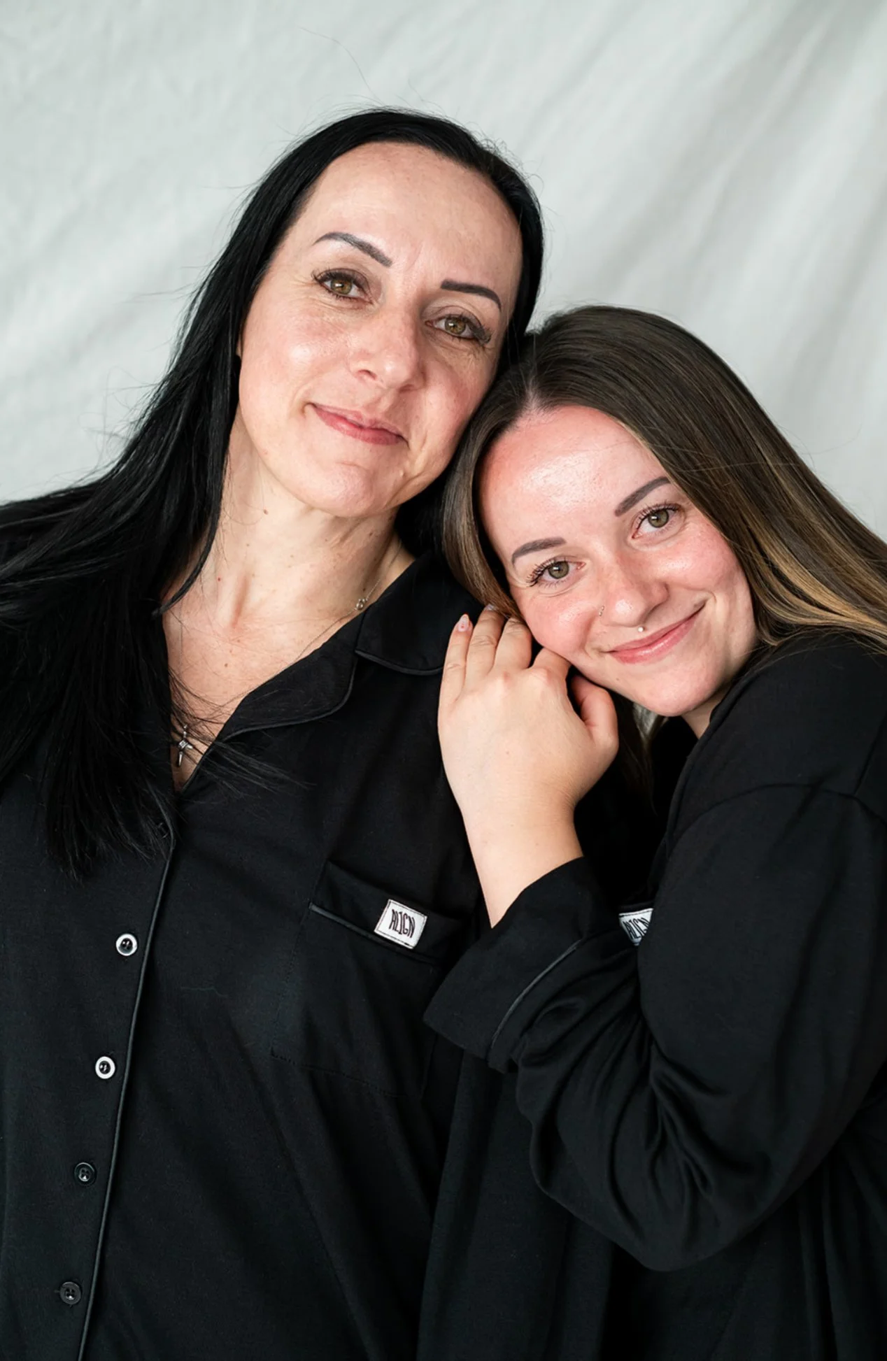 Two women, one with long black hair and the other with long brown hair, close together, both smiling softly, wearing black shirts, with a light-colored background.