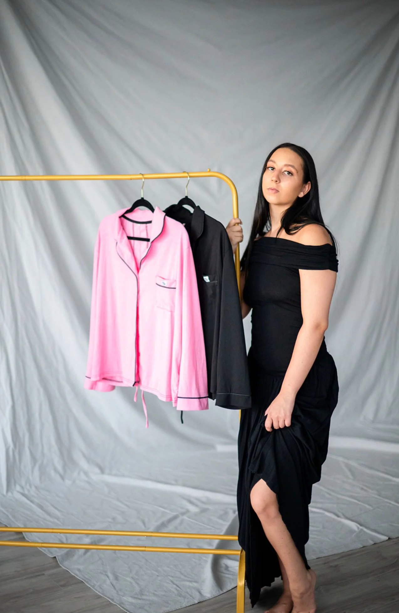 Woman in black clothing standing next to a gold clothing rack holding pink and black pajamas against a plain backdrop.