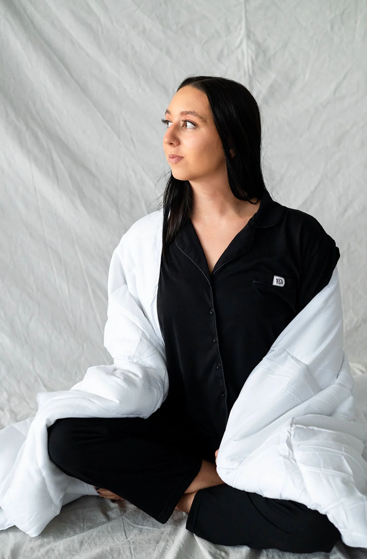 A woman with long black hair wearing black pajamas and a white jacket, sitting cross-legged and looking to her left against a gray crinkled backdrop.