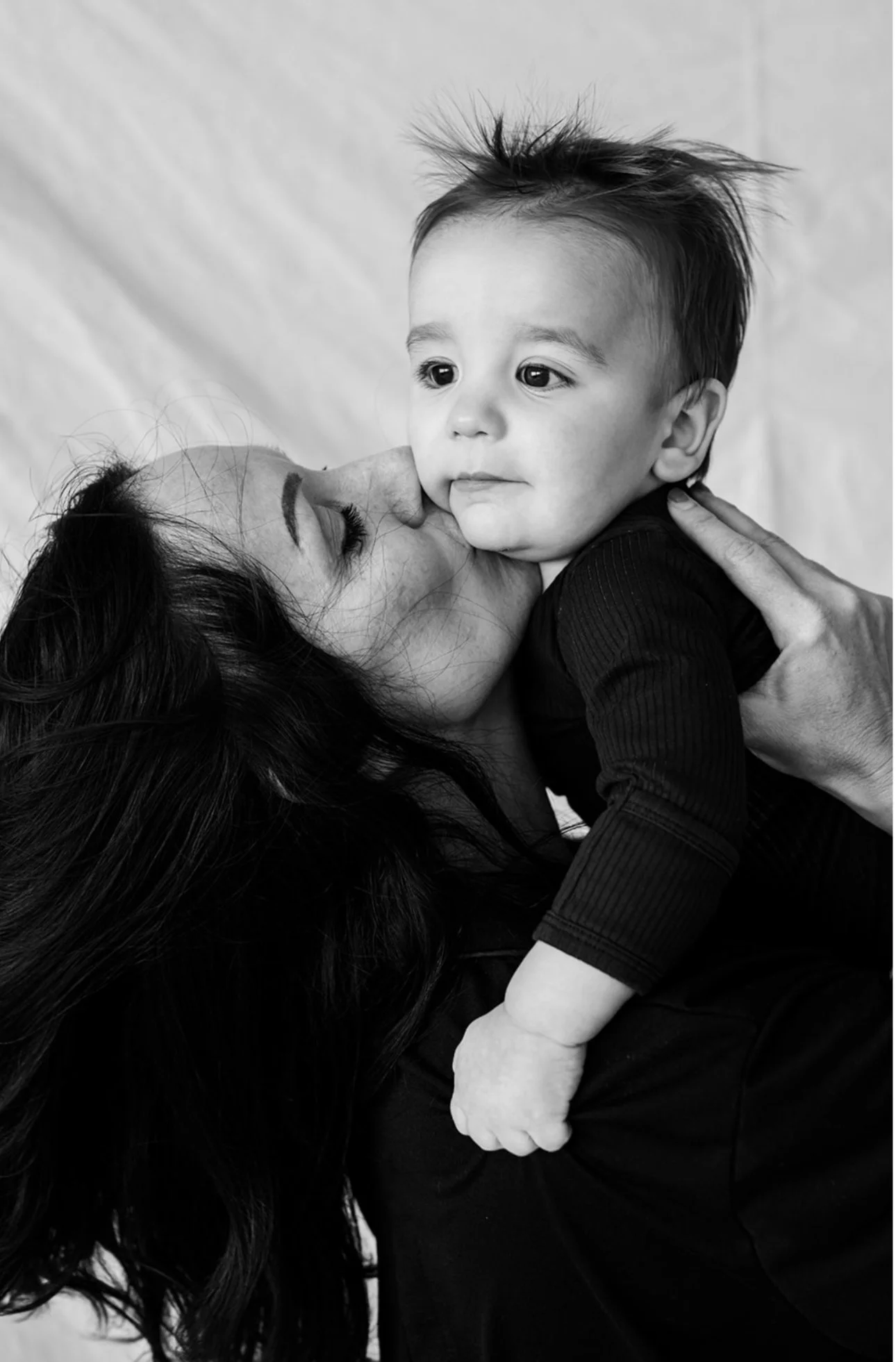 A woman with long dark hair holding a young boy, giving him a kiss on the cheek. The boy has short, spiky hair and is looking slightly to the right, while the woman is kissing his cheek. The photo is in black and white.