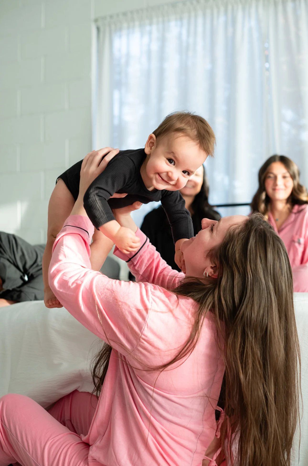 A woman in pink pajamas playing with a smiling toddler boy while other women look on in the background.