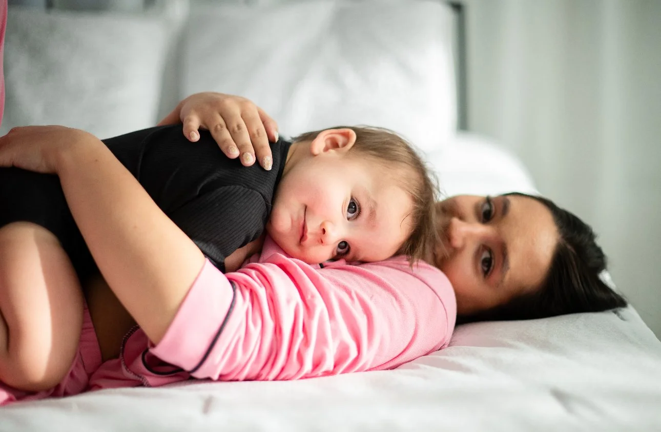 A mother lying on her side in bed holding her young child, who is lying on her back with his head resting on her shoulder, both looking at the camera.