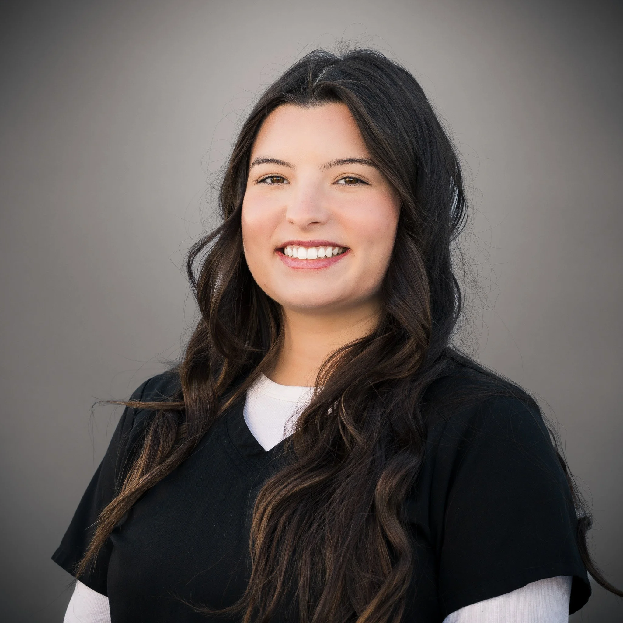 Smiling person with long brown hair standing outdoors, surrounded by foliage and a metal fence in the background.