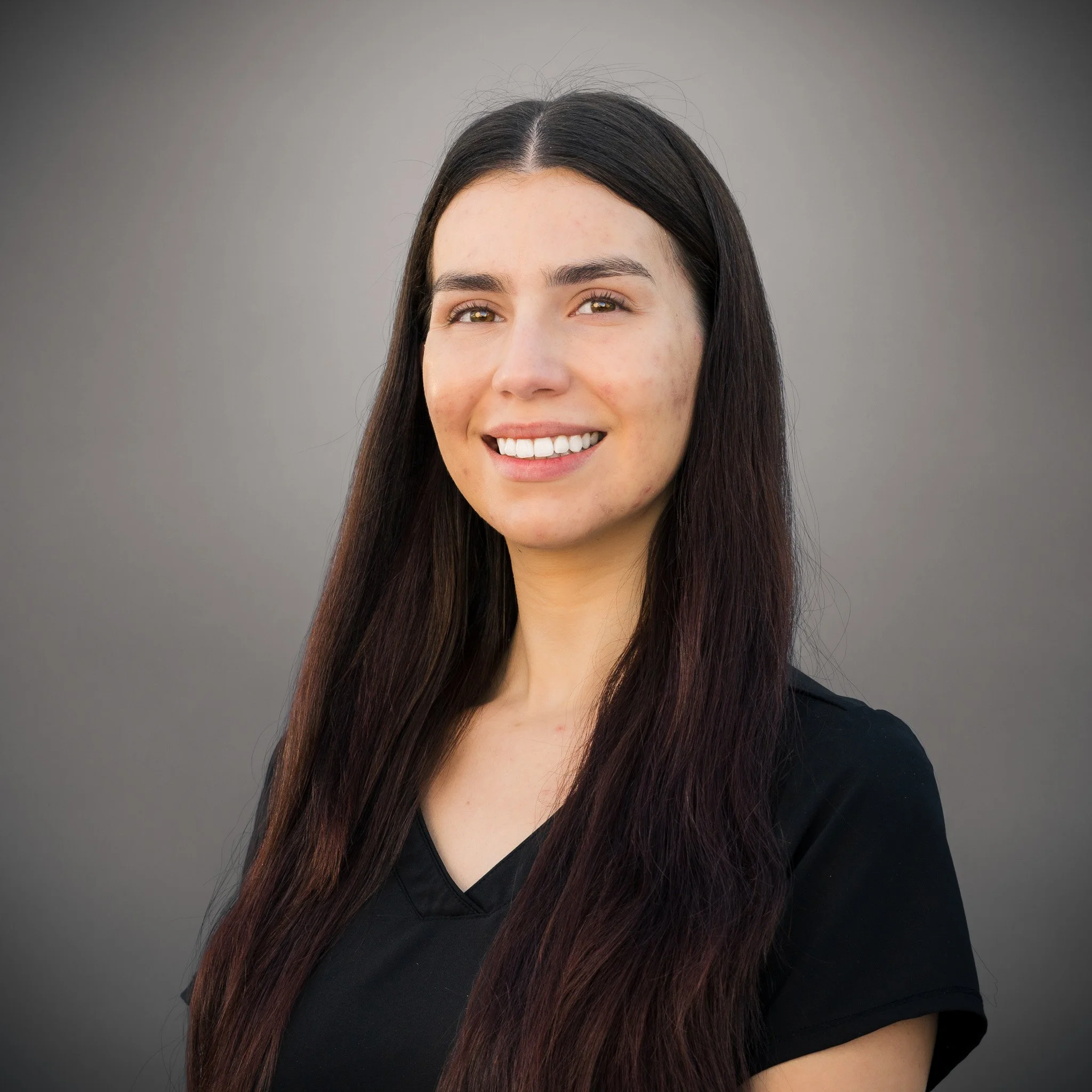 Woman smiling in front of a stone wall, wearing a dark red top, with long dark hair.