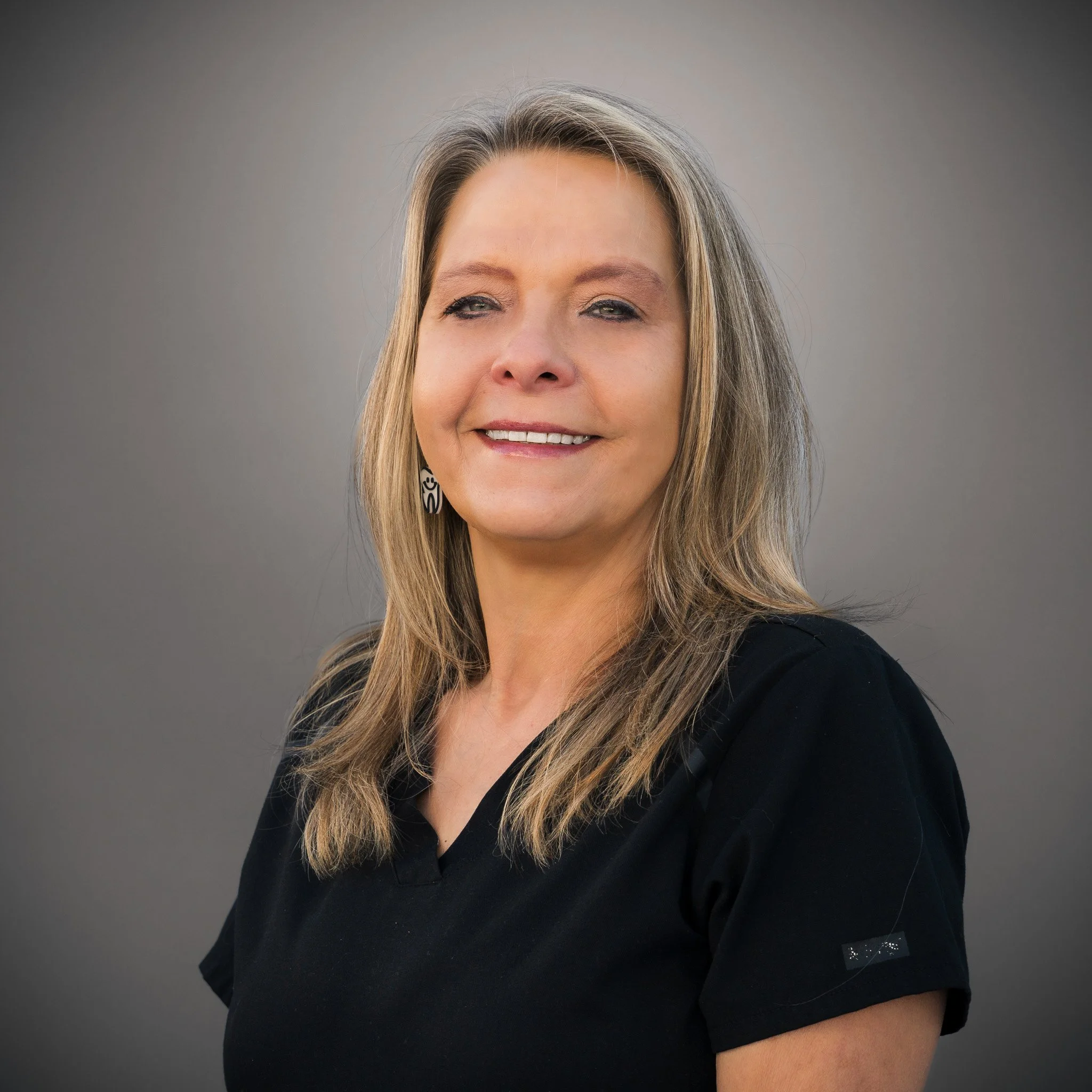 Woman with blond curly hair smiling outdoors, background of foliage and bare branches.