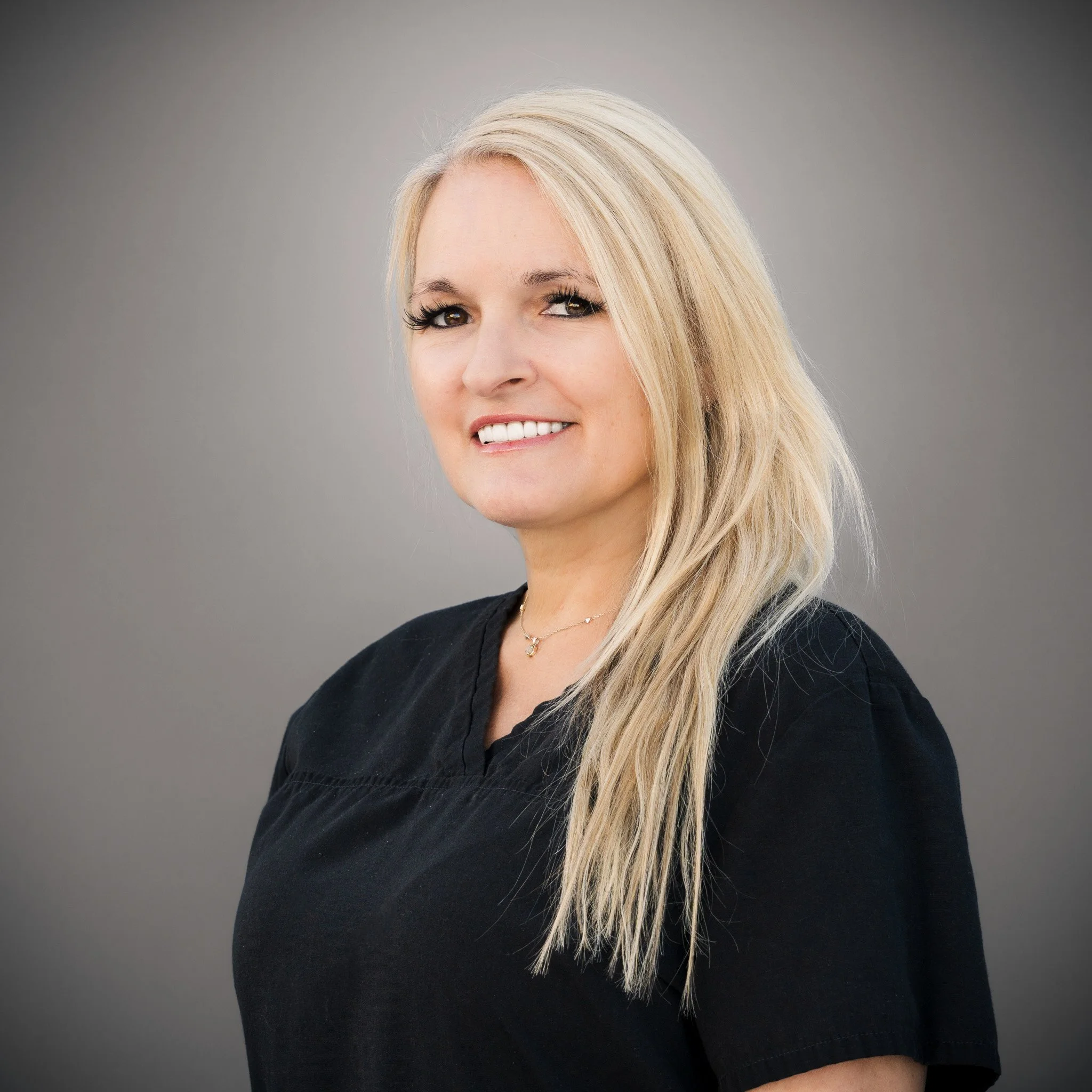Smiling woman with braided blonde hair wearing a black top and necklace, standing in front of a stone wall.