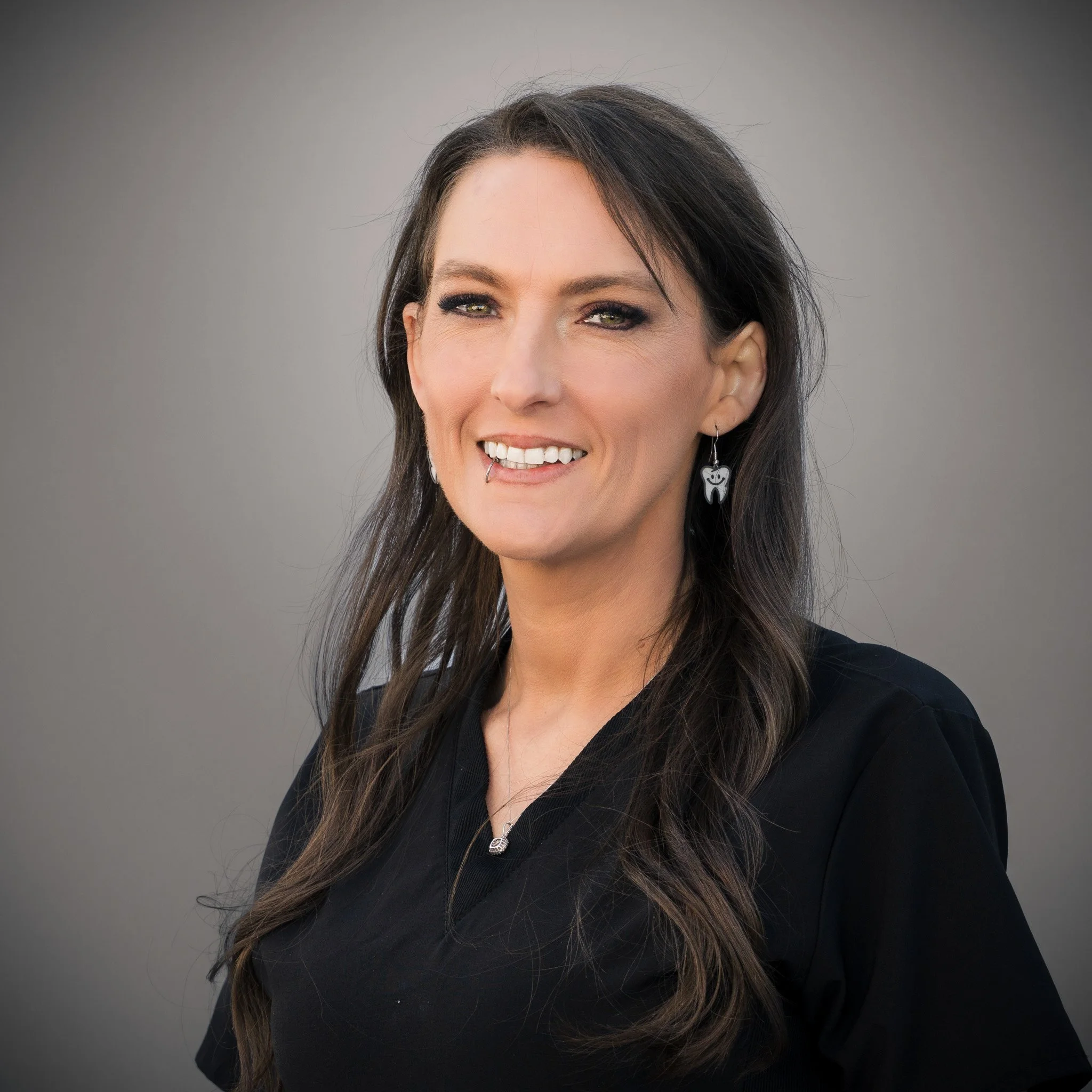 Woman with a lip piercing and braided hair, wearing a black top and necklace, standing in front of a stone wall.