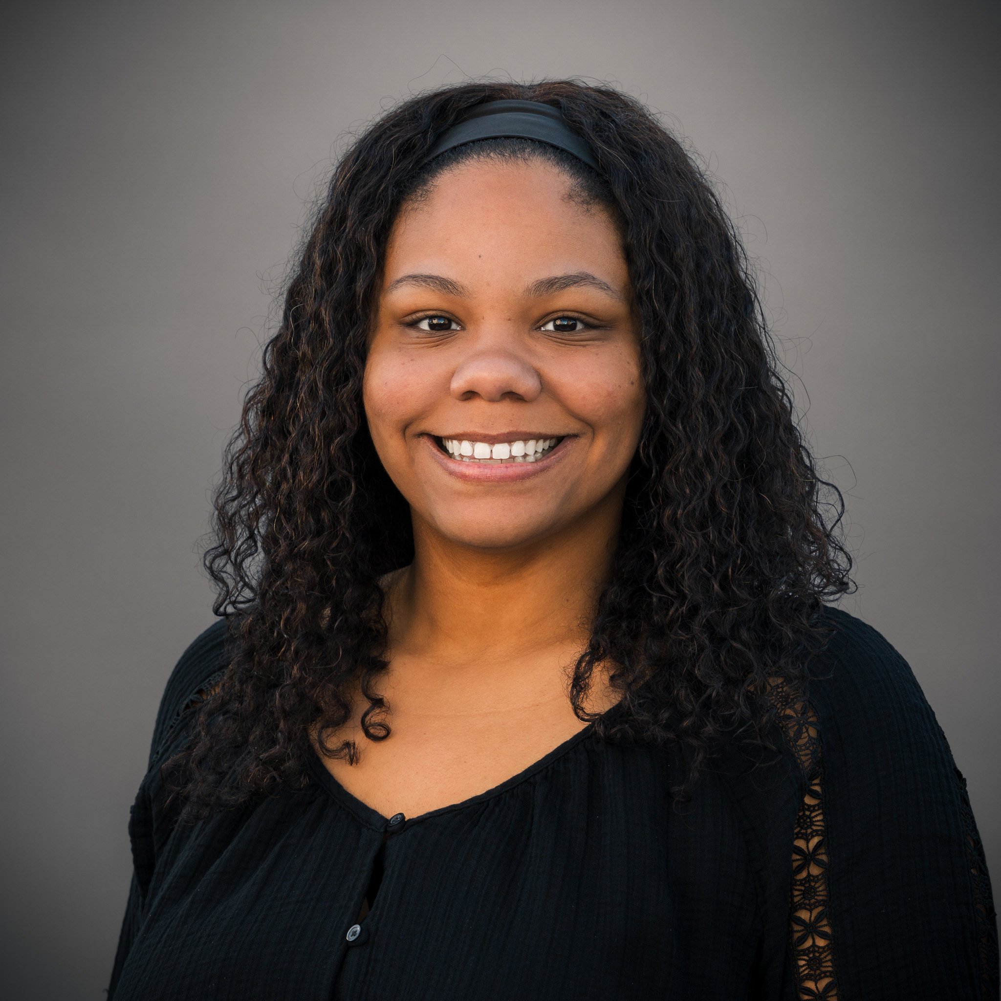 Smiling person in a black blazer outdoors with a natural background.