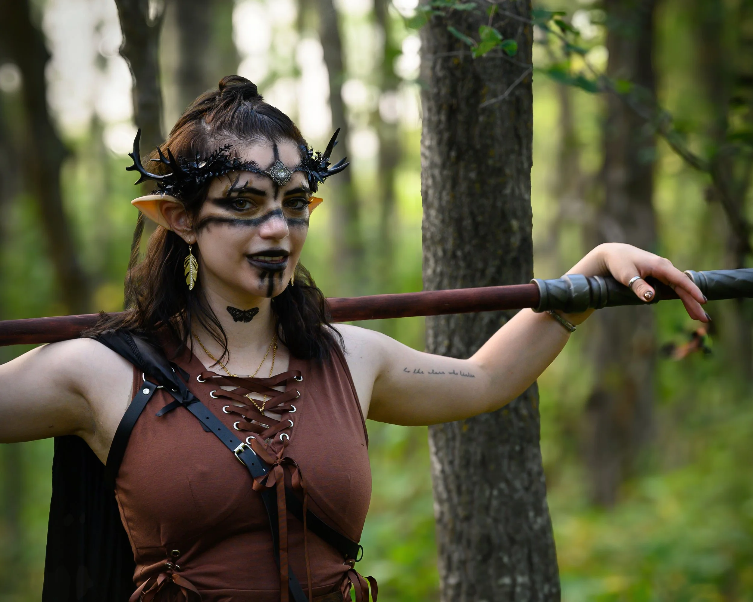 Fantasy image of a woodland fae holding a spear over her shoulders, arms draped across it. She has a brown tied sleevless tunic and tattooos on her neck and arms. The image is in a forest outside Edmonton.