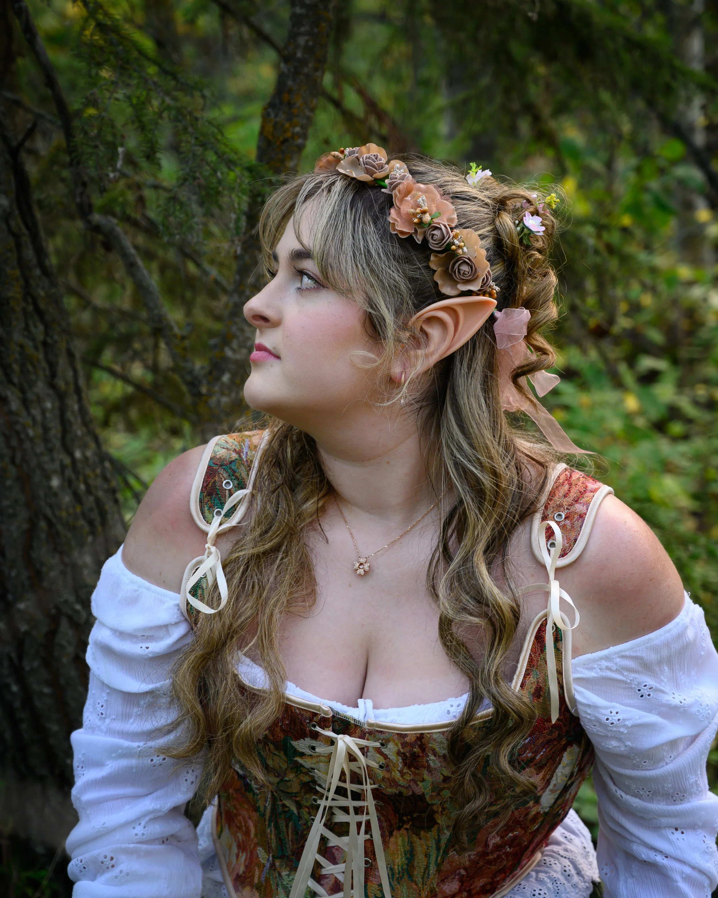 Fantasy photo taken in a forest outside of St. Albert of a fae woman with pointed ears seated on a log. She has a flower crown and flowing golden hair.