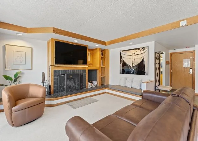 Living room with brown leather sofa, tan armchair, fireplace, wall-mounted TV, built-in shelves, artwork, and a small seating nook near the entrance.