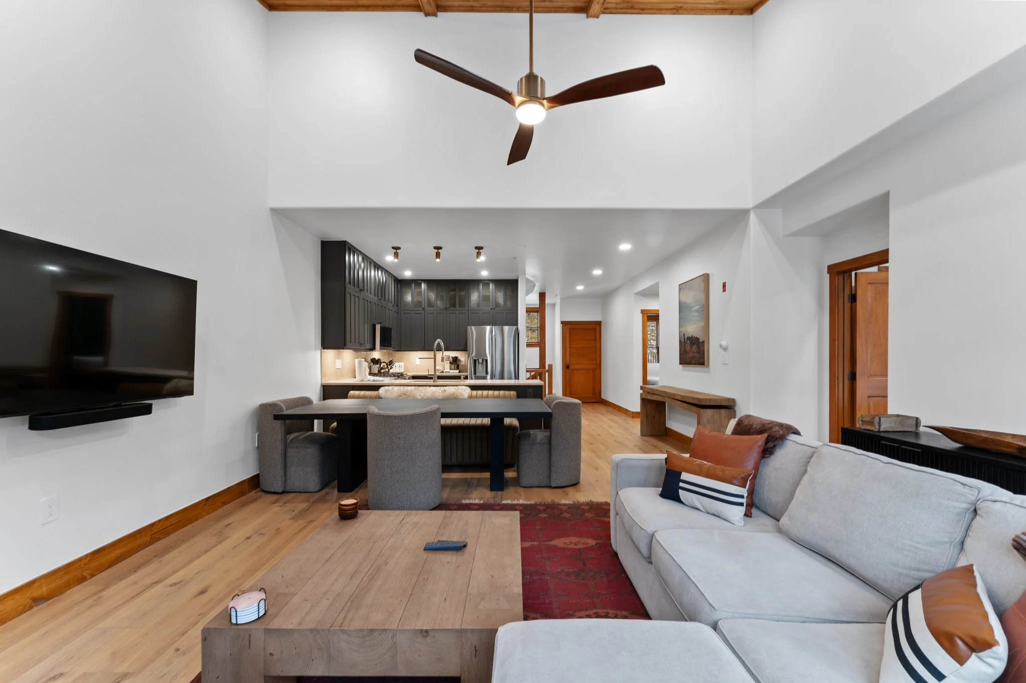 Living room with white walls, wooden floor, a large flat-screen TV mounted on the wall, a beige sectional sofa with pillows, a wooden coffee table, a modern ceiling fan, and a view into the kitchen with dark cabinets, stainless steel refrigerator, an