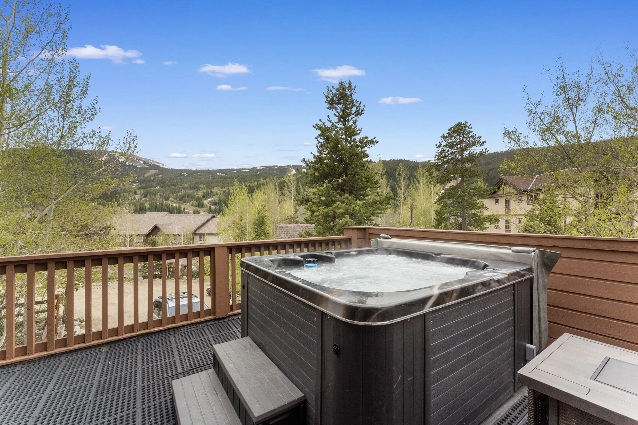 Outdoor hot tub on a wooden deck overlooking a mountainous landscape with trees and houses under a blue sky.