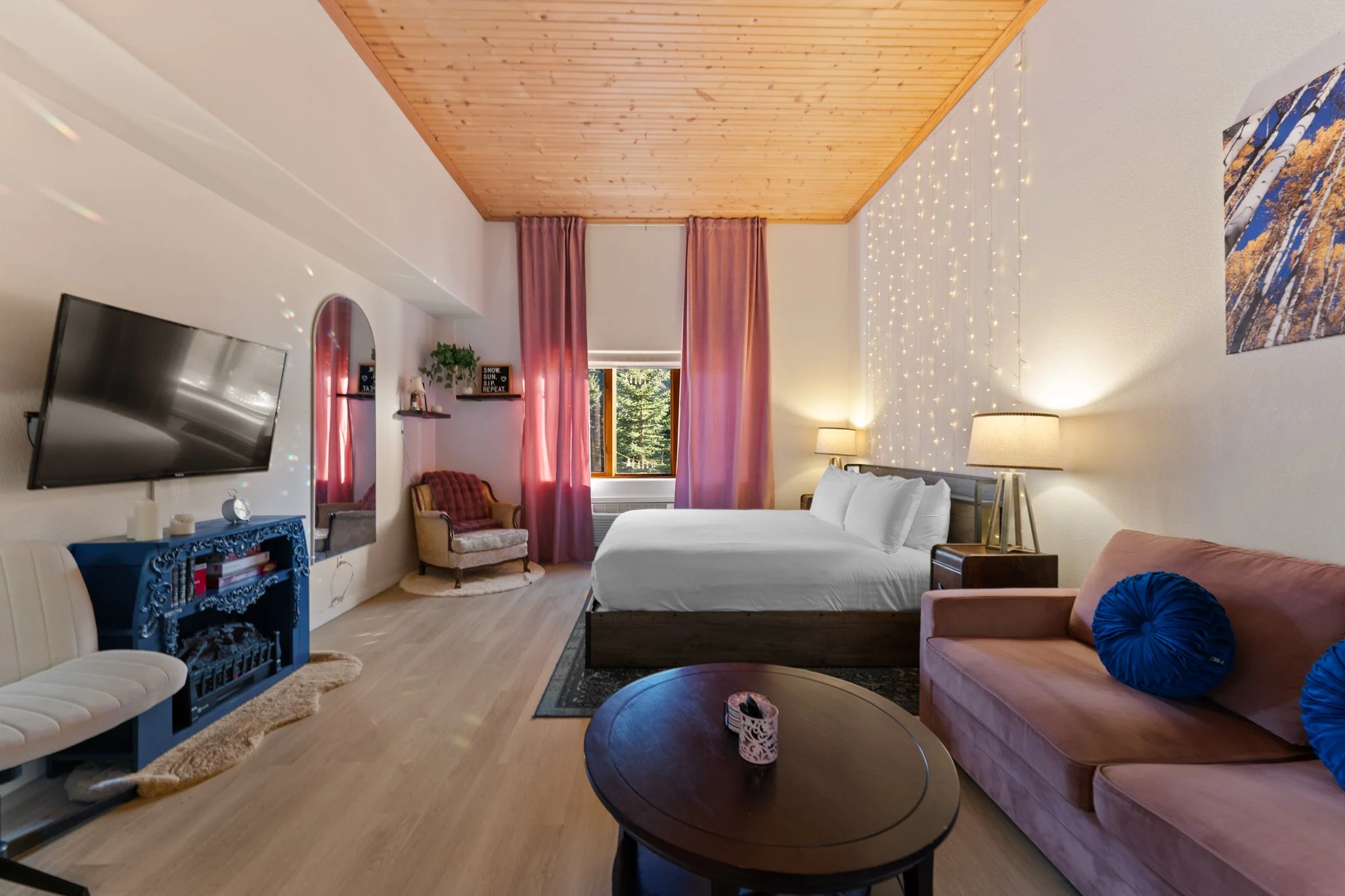 A cozy hotel room with a wooden ceiling, white walls, a large bed, a pink sofa with blue pillows, a round coffee table, a wall-mounted TV, pink curtains, fairy lights, lamps, a vintage armchair, and a window showing trees outside.