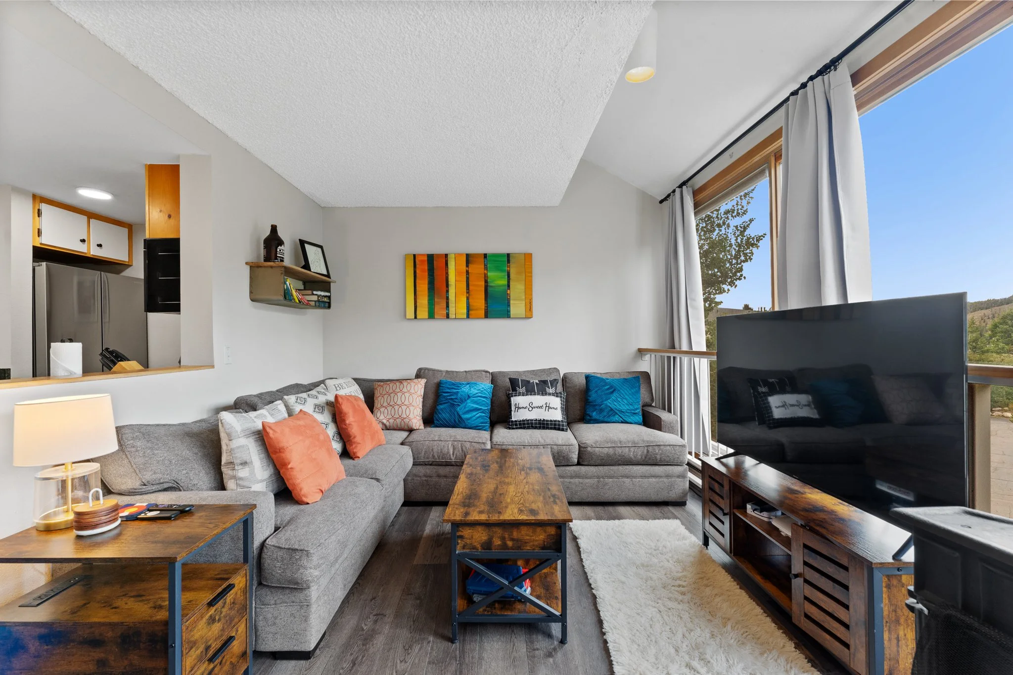 Living room with gray sectional sofa, colorful throw pillows, wooden coffee table, large flat-screen TV, and a large window with white curtains showing a scenic outdoor view