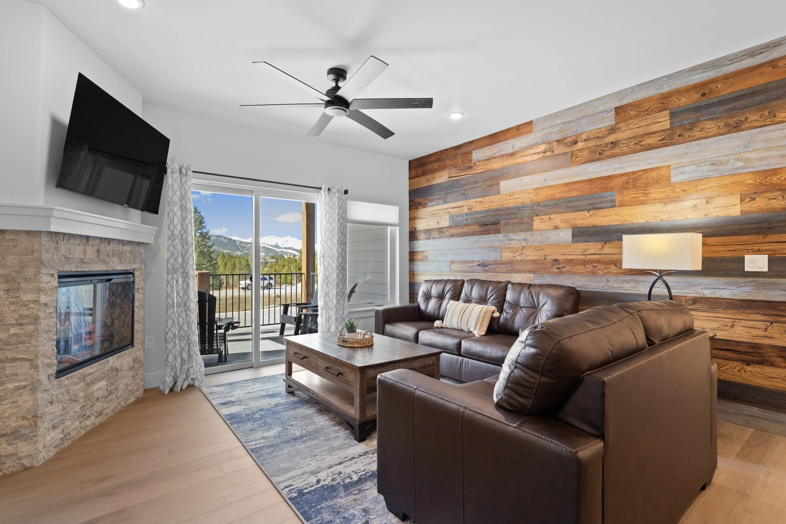 Living room with brown leather sofas, wooden coffee table, stone fireplace, large window with mountain view, sliding glass door with curtain leading to balcony, wooden accent wall, ceiling fan, lampshade, and mountain scenery outside.
