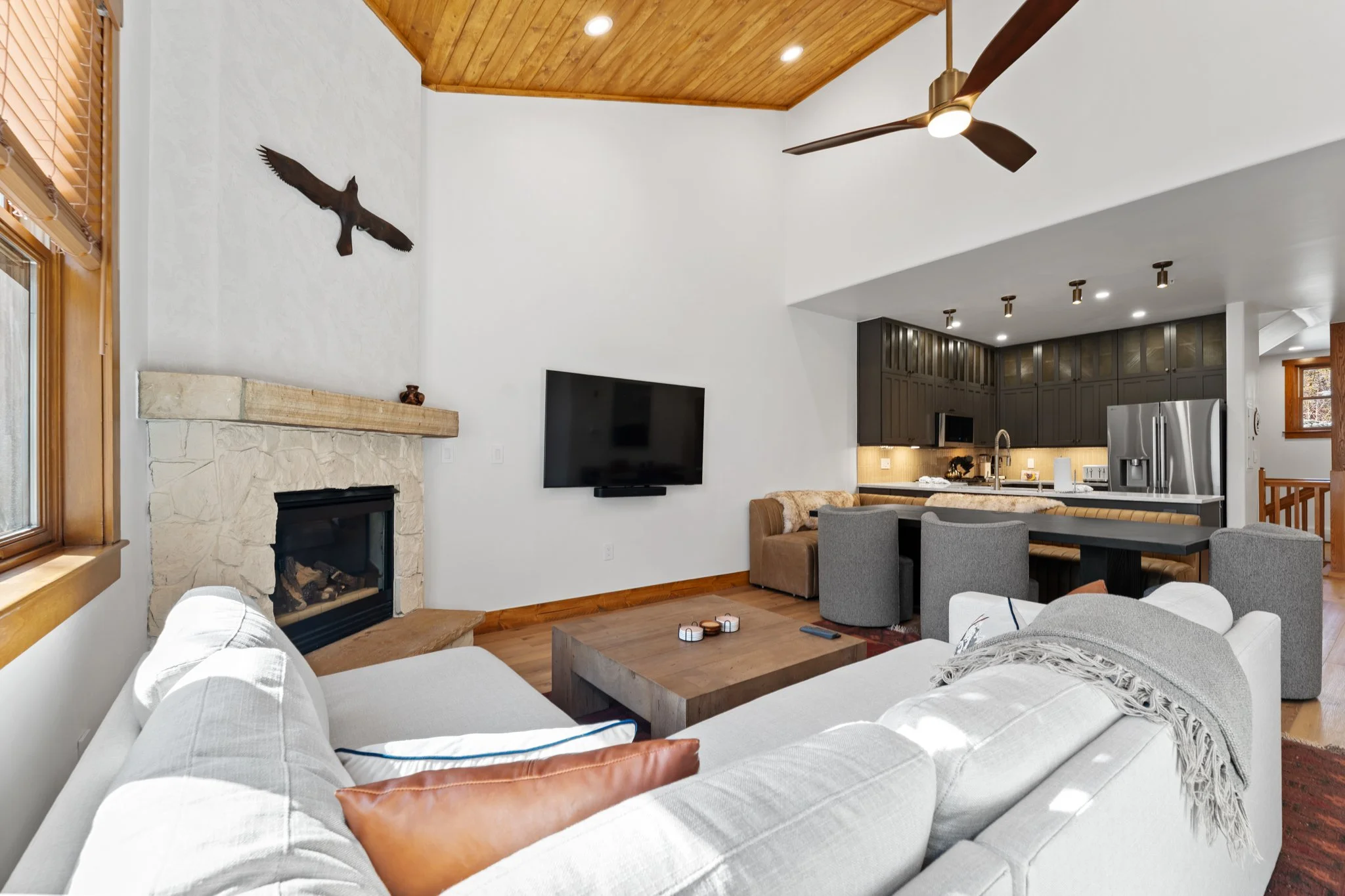 Open-concept living room with white sofa, stone fireplace, wall-mounted television, and a kitchen with dark cabinets and stainless steel appliances.