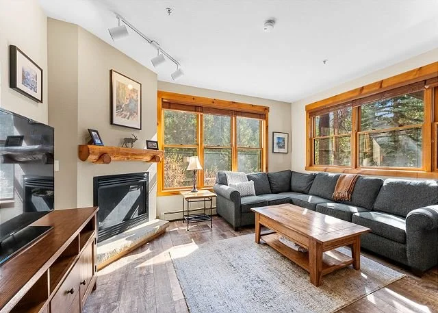 Living room with large windows, gray sectional sofa, wooden coffee table, fireplace with wooden mantel, TV stand, lamp, framed pictures on the wall, and a beige rug.