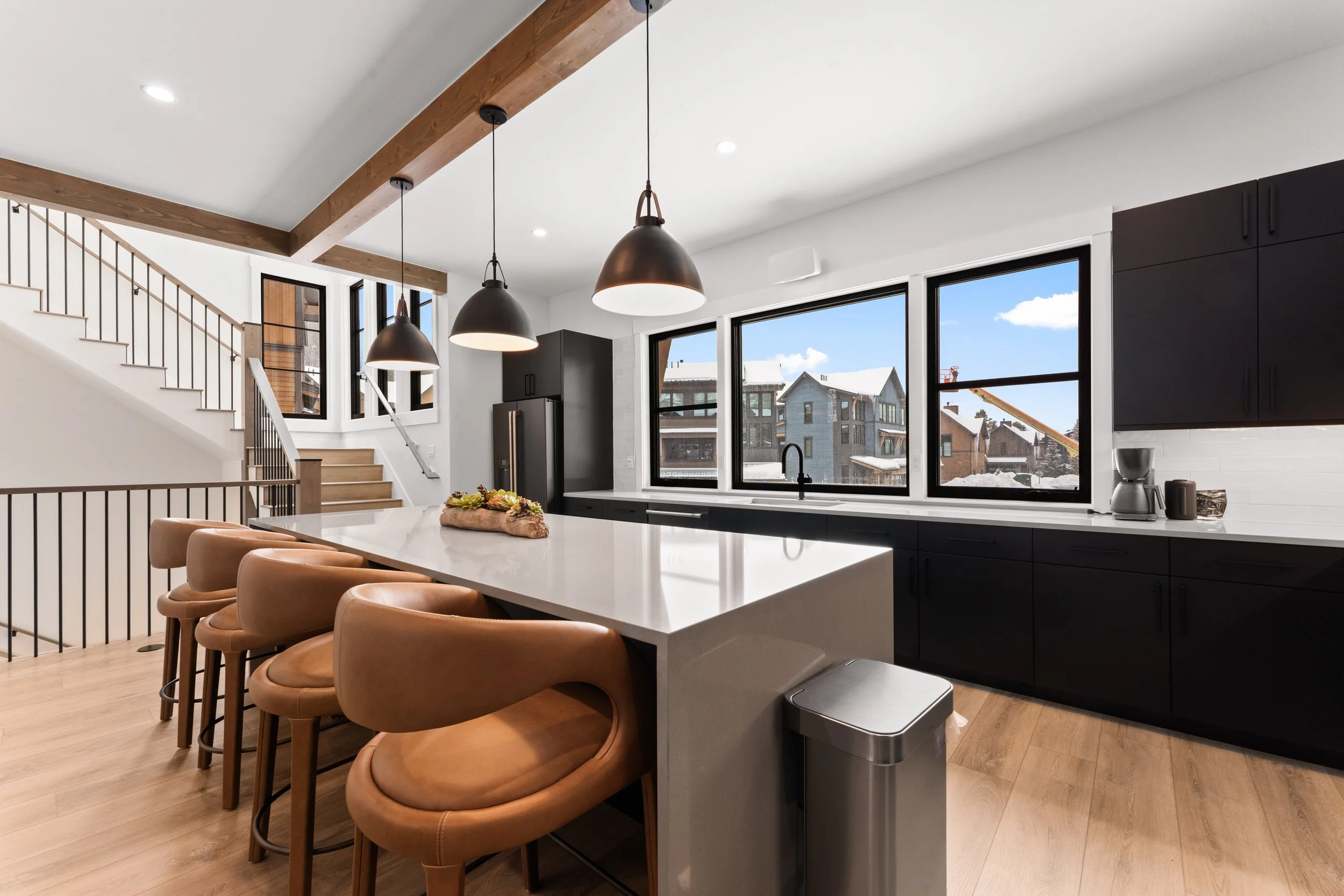 Modern kitchen with black cabinetry, large window, white countertops, hanging pendant lights, and beige bar stools at an island.