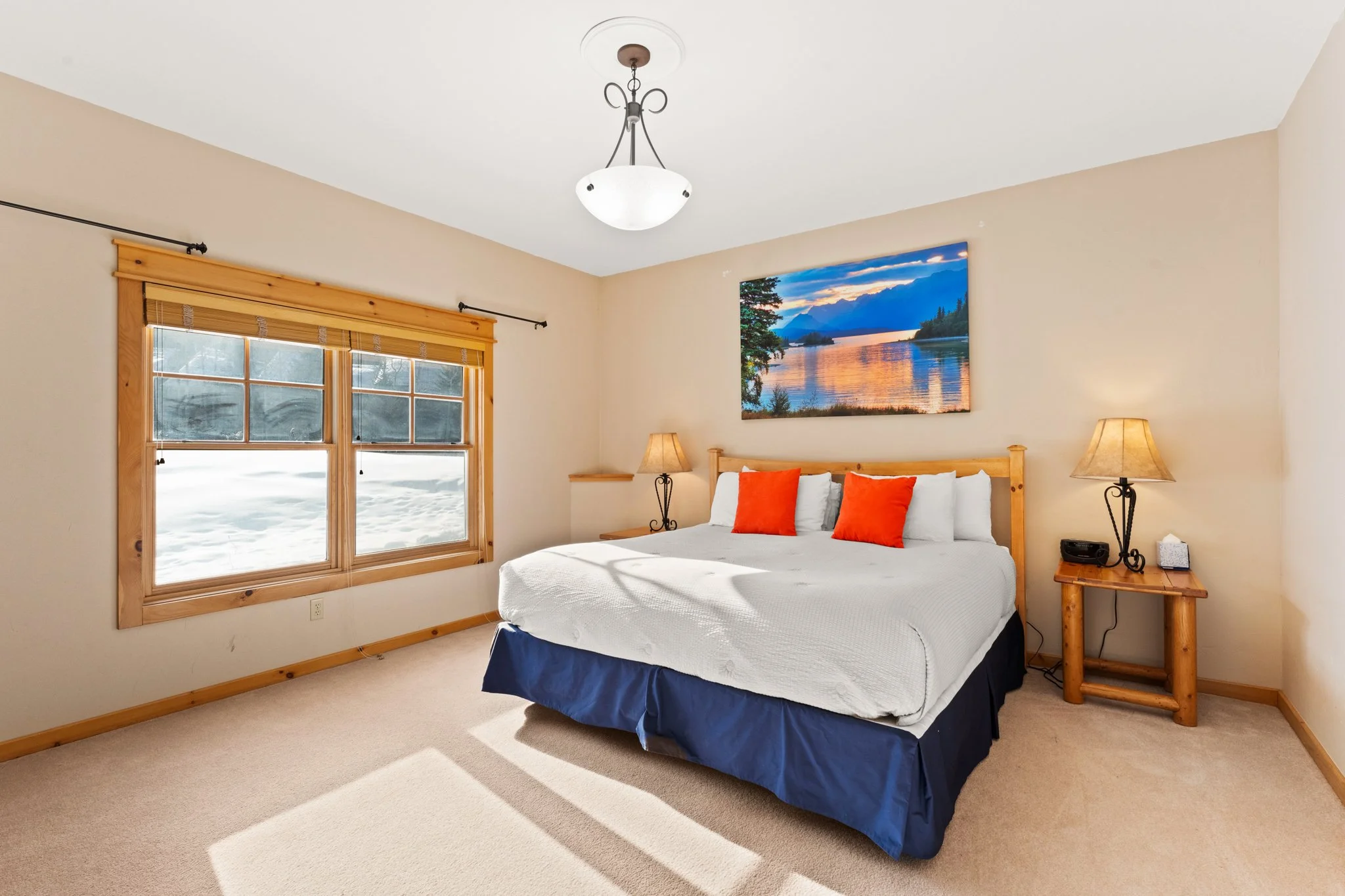 Bedroom with beige walls, wooden window trim, a bed with white bedding, three red pillows, and a blue bed skirt. Two wooden nightstands with lamps, a landscape painting above the bed, and a window revealing snow outside.