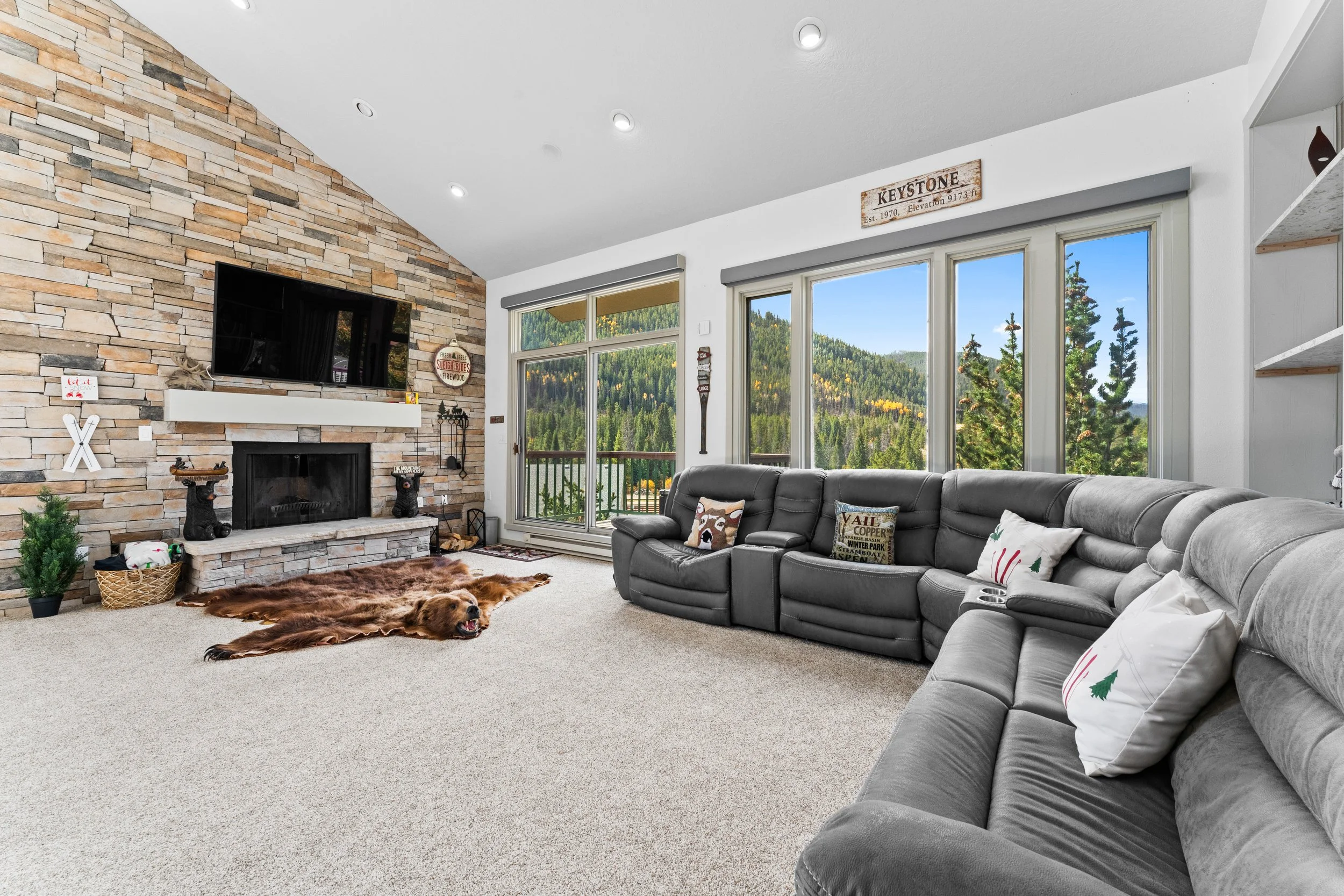 Living room with a stone fireplace, a gray sectional sofa, large windows with mountain view, and dog lying on a fur rug.