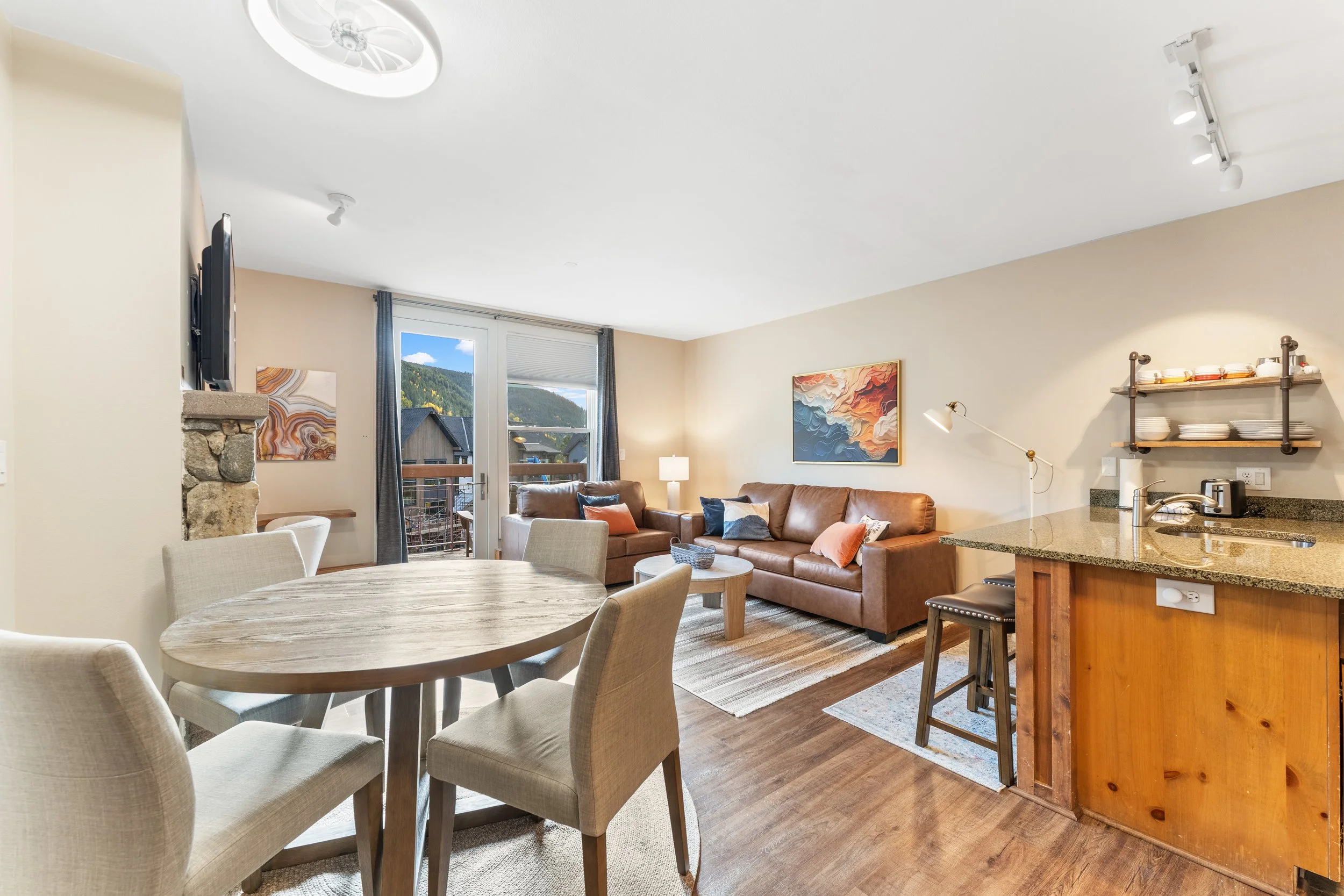 Living room with brown leather sofa, armchair, artwork, and large windows showing mountain views, adjacent to a kitchen with granite countertop and open shelving.
