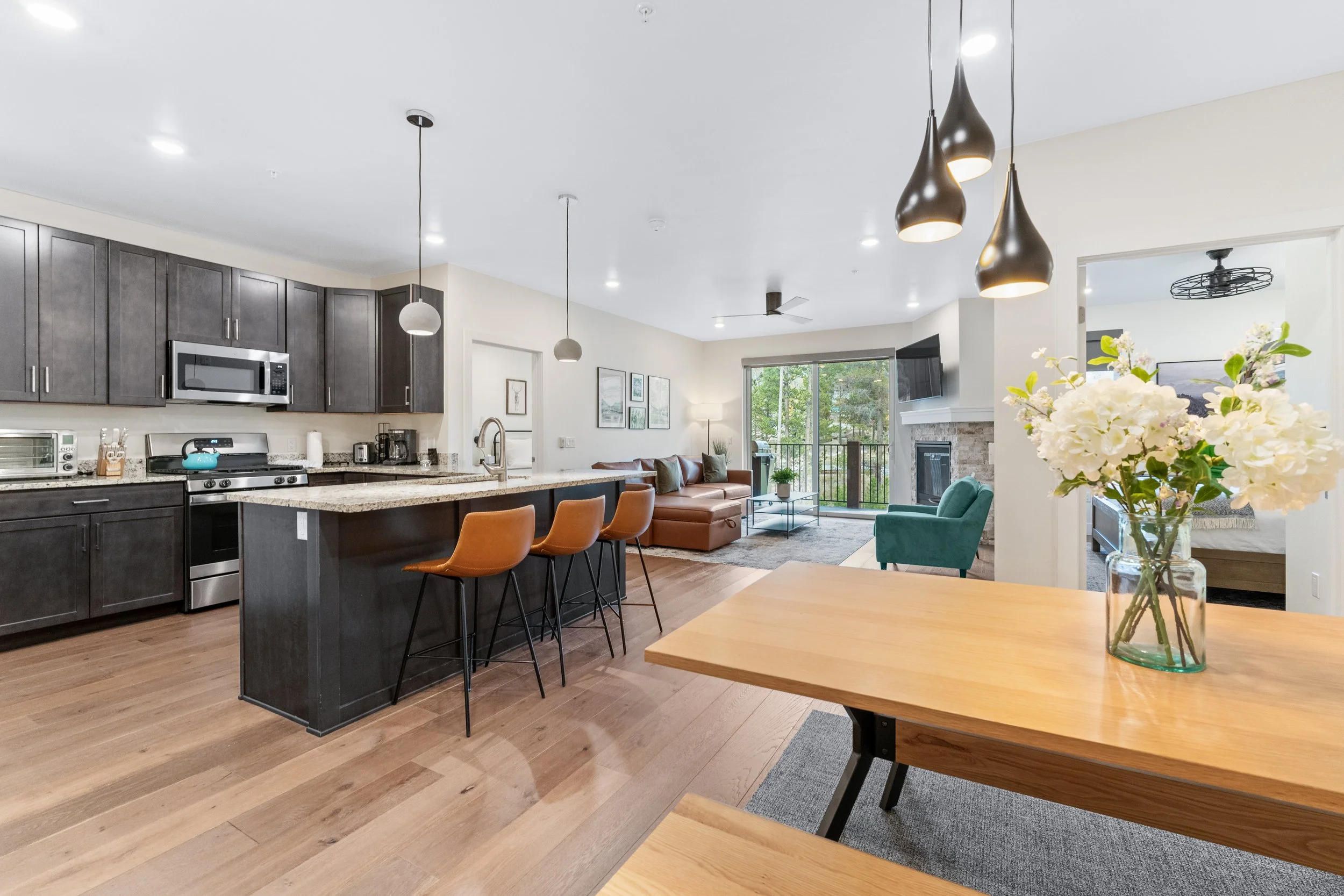 Open-concept kitchen and living room with dark cabinets, granite countertops, a dining area with a wooden table and a vase of white flowers, a kitchen island with three brown chairs, and a living room with leather and upholstered sofas, a fireplace, 