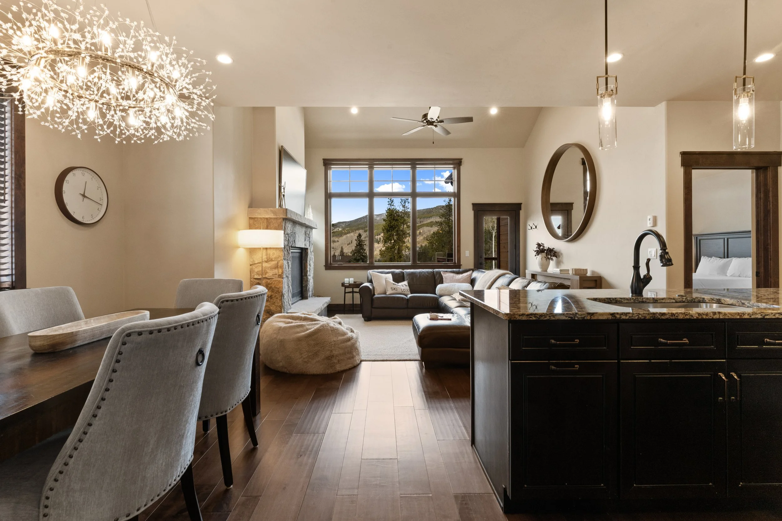 Open-concept living room and dining area with large window showing mountains and trees, fireplace, beige walls, modern chandelier, beige chairs, dark brown hardwood floors, and kitchen island with black cabinets and granite countertop.